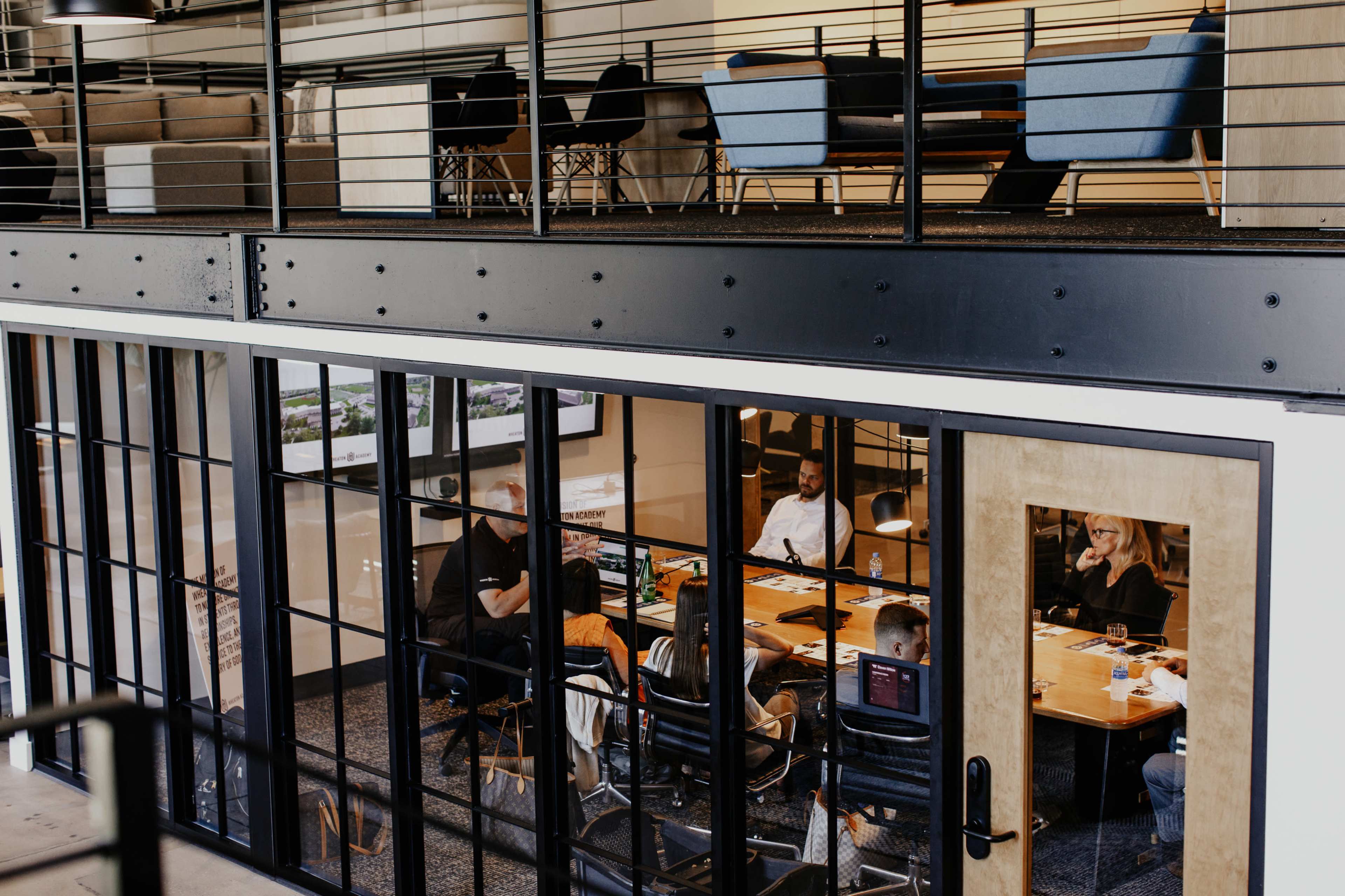 A group of people is having a meeting around a table in a conference room, while additional seating is visible on an upper level.