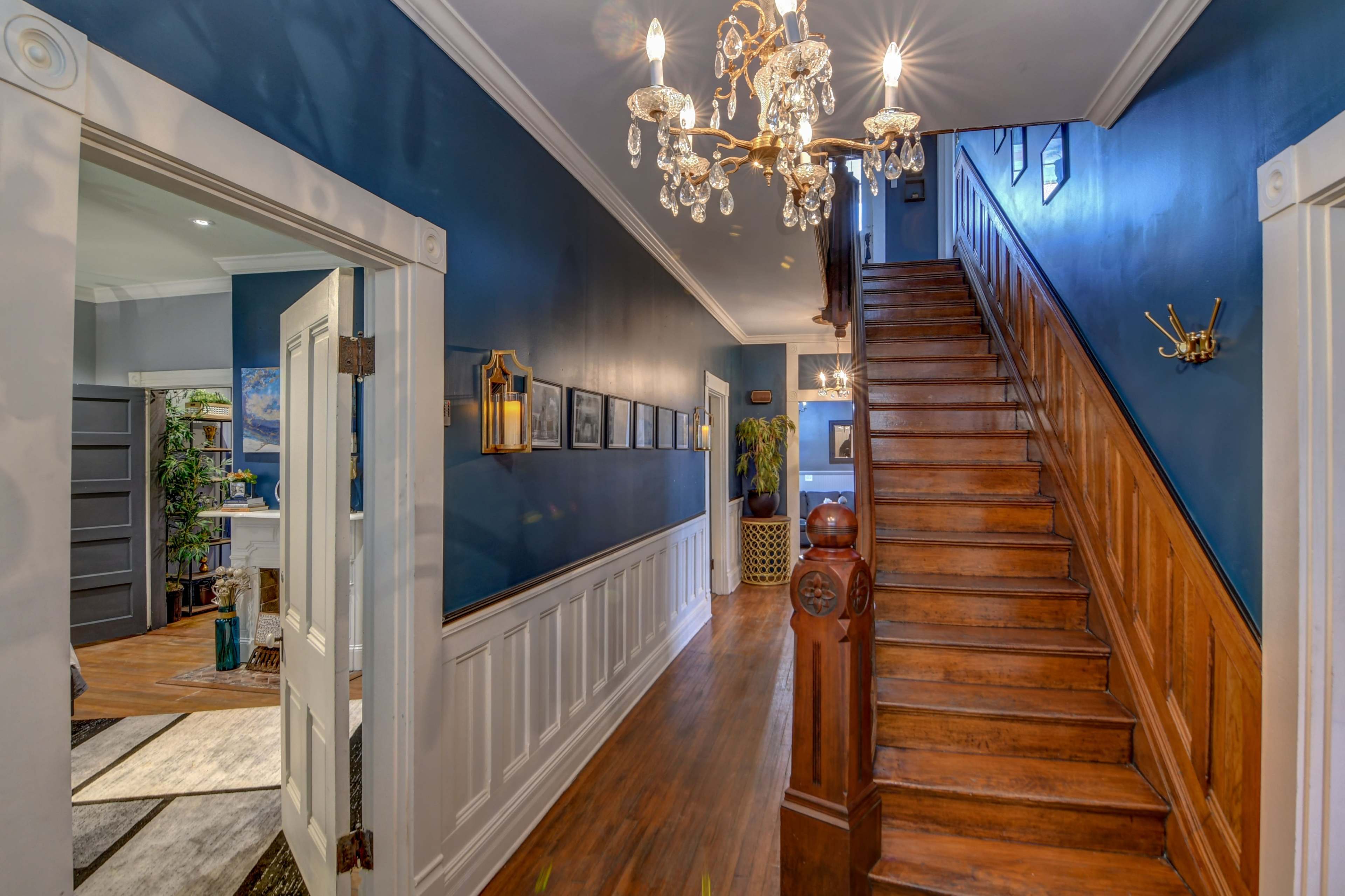 A well-lit hallway features a wooden staircase, blue walls, and a chandelier, with framed pictures lining the walls.