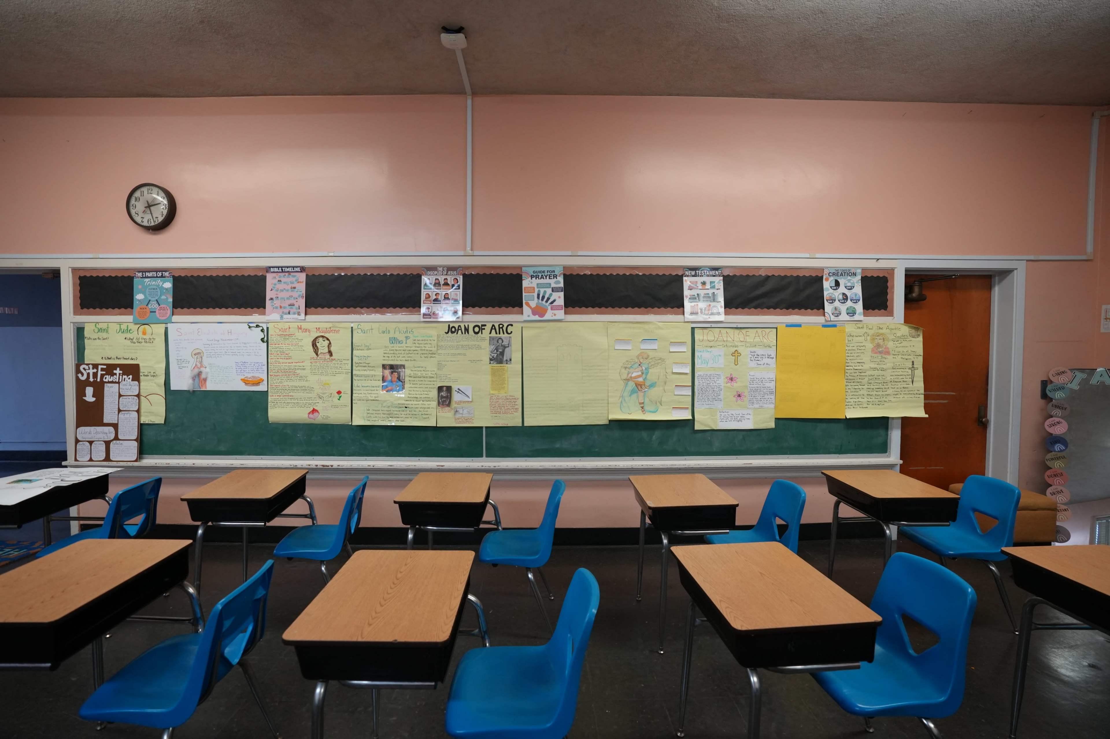 The image shows a classroom with empty blue chairs and wooden desks facing a green chalkboard and a bulletin board covered with various posters and announcements.