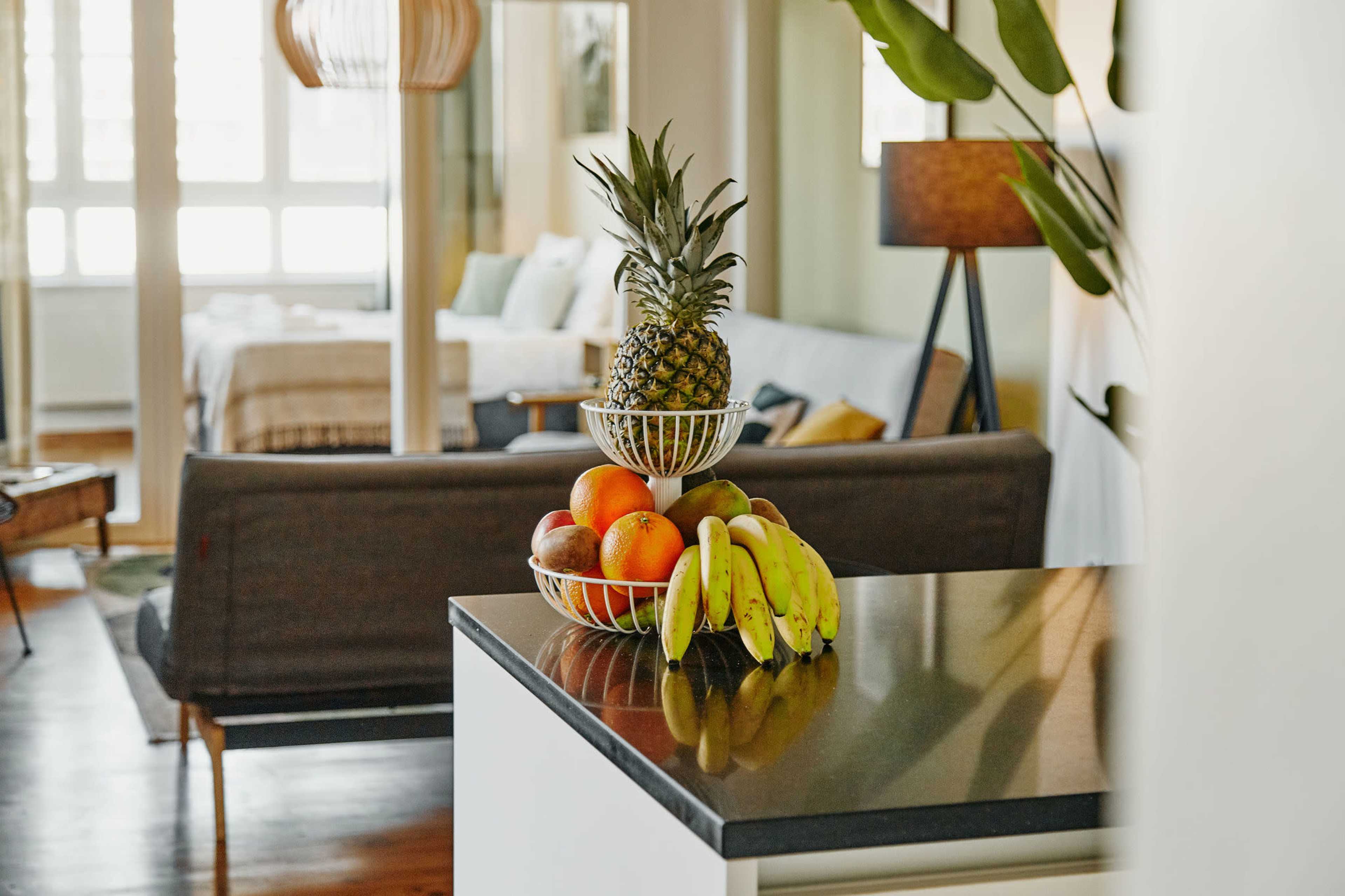 A kitchen countertop displays a bowl of assorted fruits, including a pineapple, bananas, and apples, with a living space in the background.