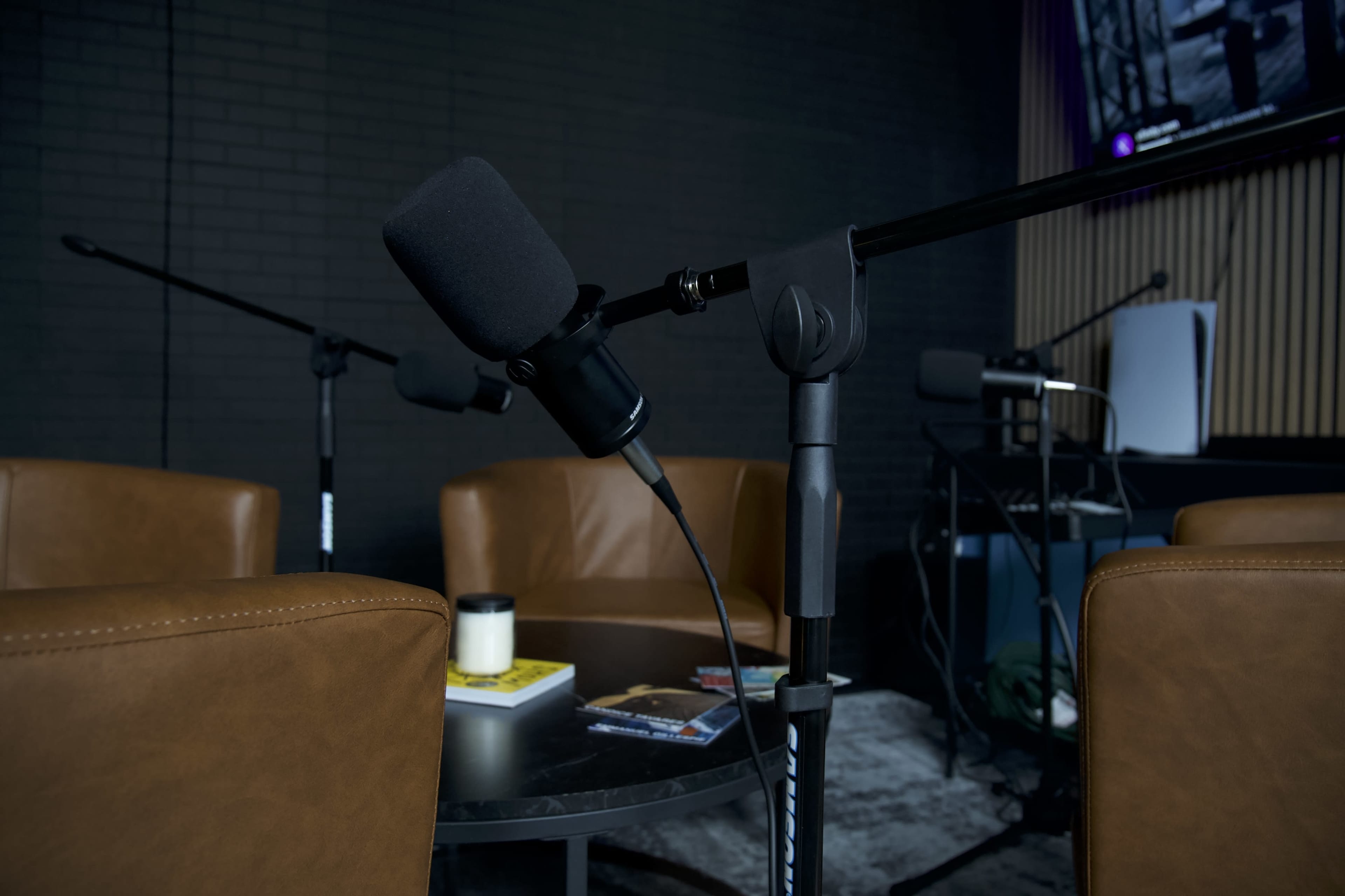 The image shows a podcast recording setup with microphones positioned near brown leather chairs and a coffee table scattered with books and a candle.