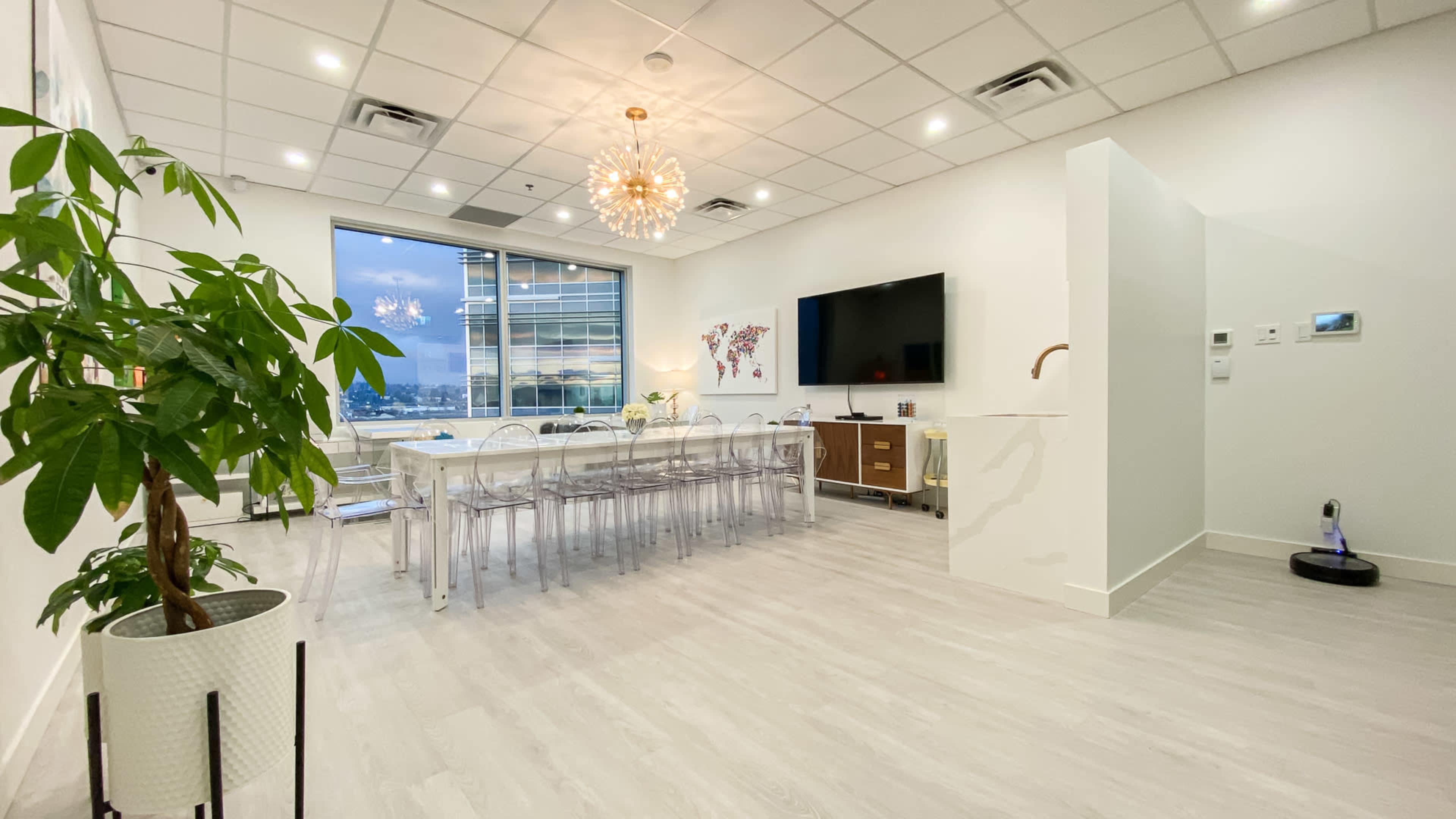 A modern conference room with a long table, transparent chairs, a large TV, and a view of the city through a window.