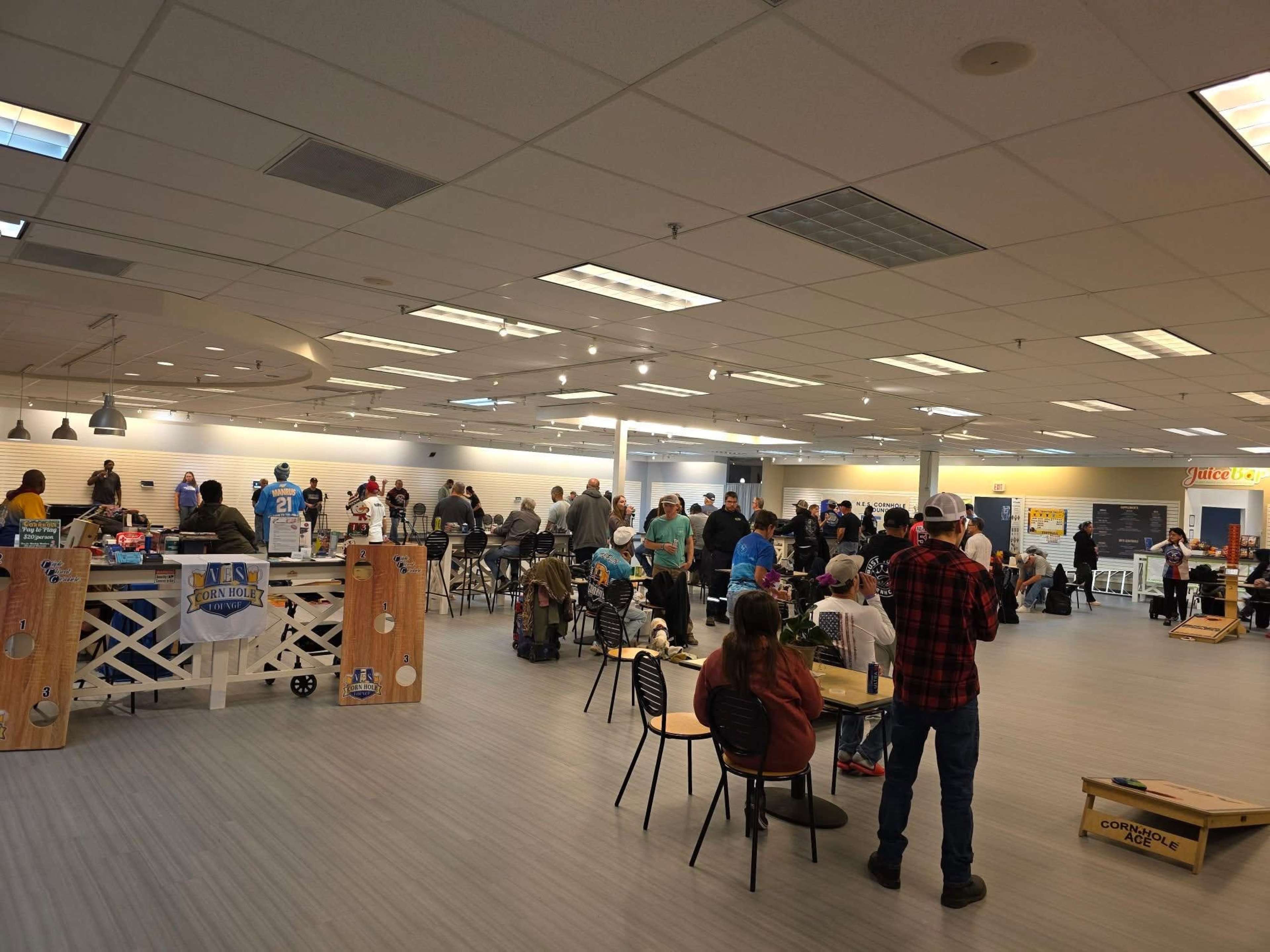 A crowded indoor space with people seated and standing, featuring tables and games, likely part of a community event or gathering.