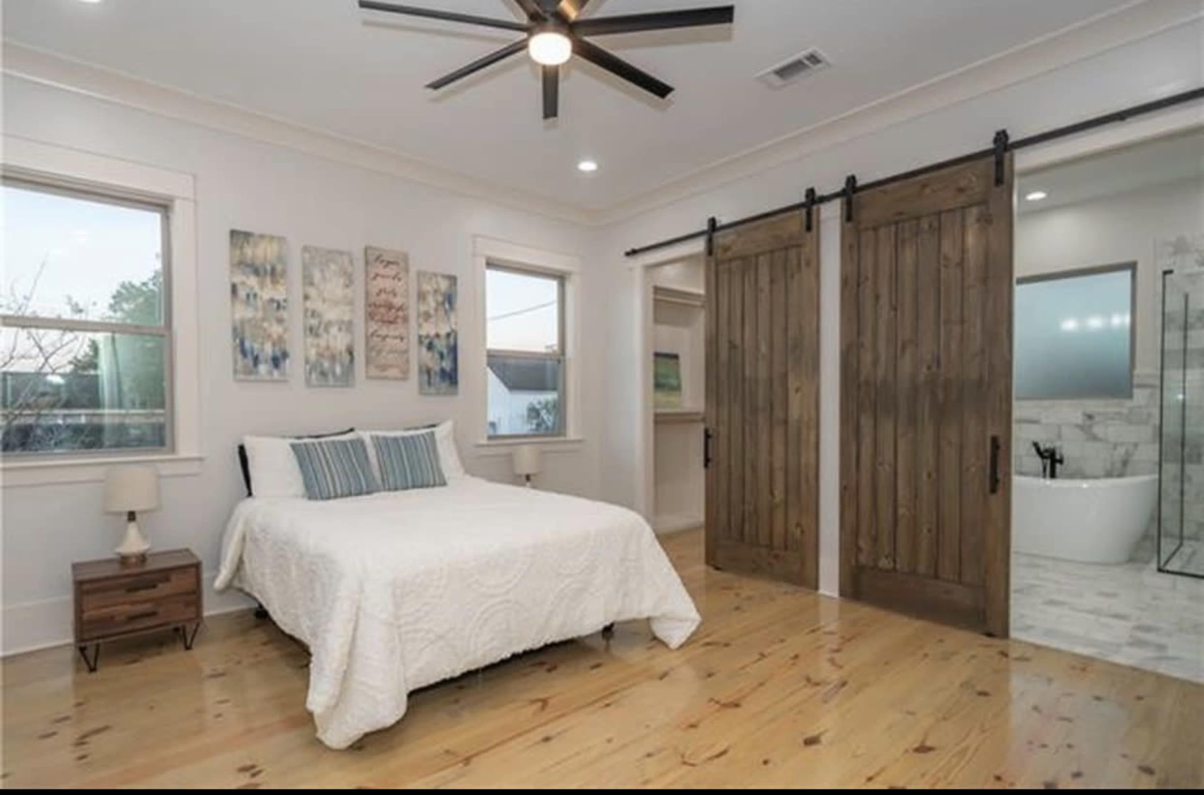 The image shows a modern bedroom with a white quilted bedspread, wooden floors, and sliding barn doors leading to a bathroom.
