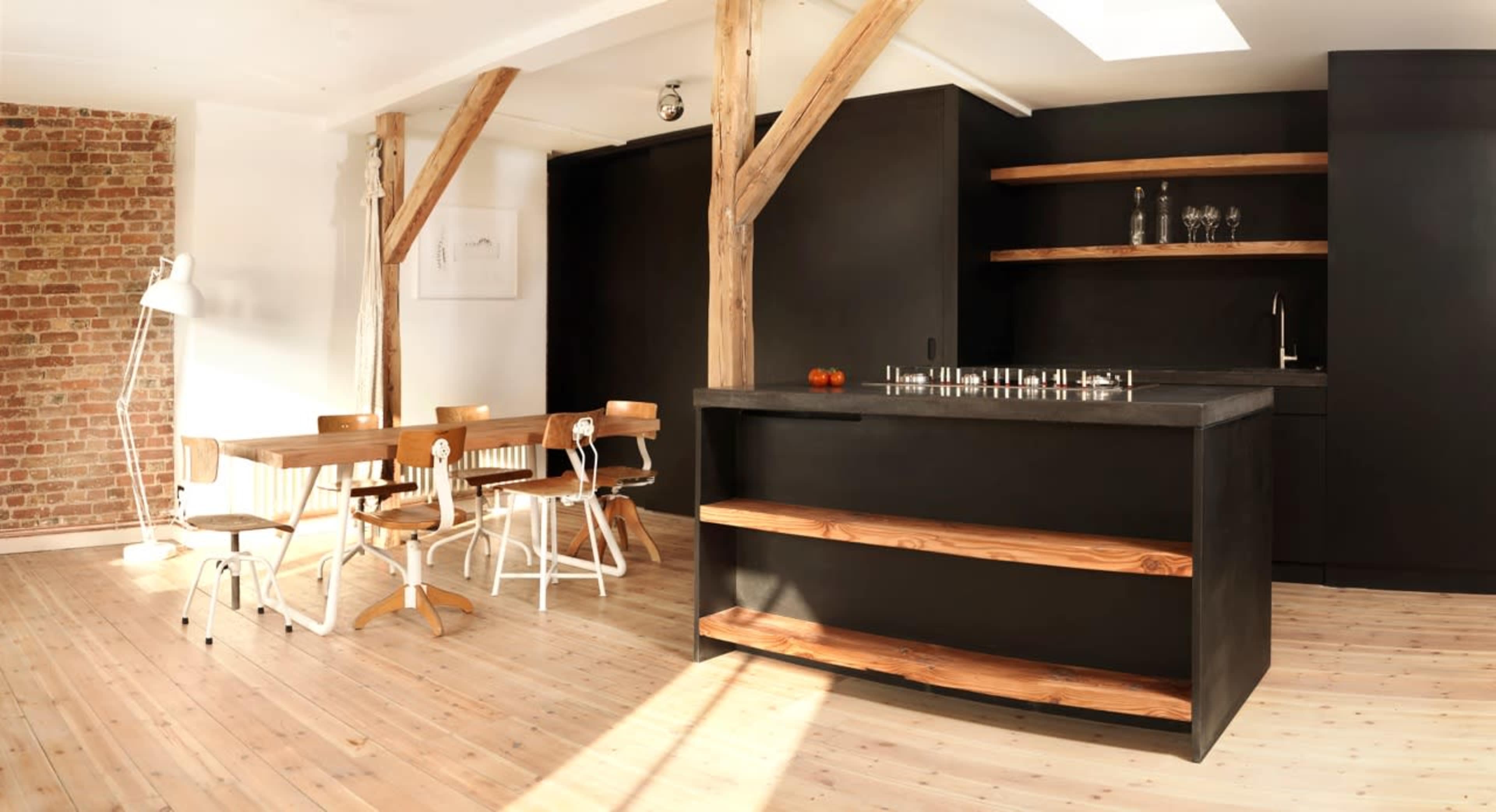 The image depicts a modern kitchen and dining area featuring a dark cabinetry, wooden shelves, a countertop with an integrated stove, and a dining table with four chairs.