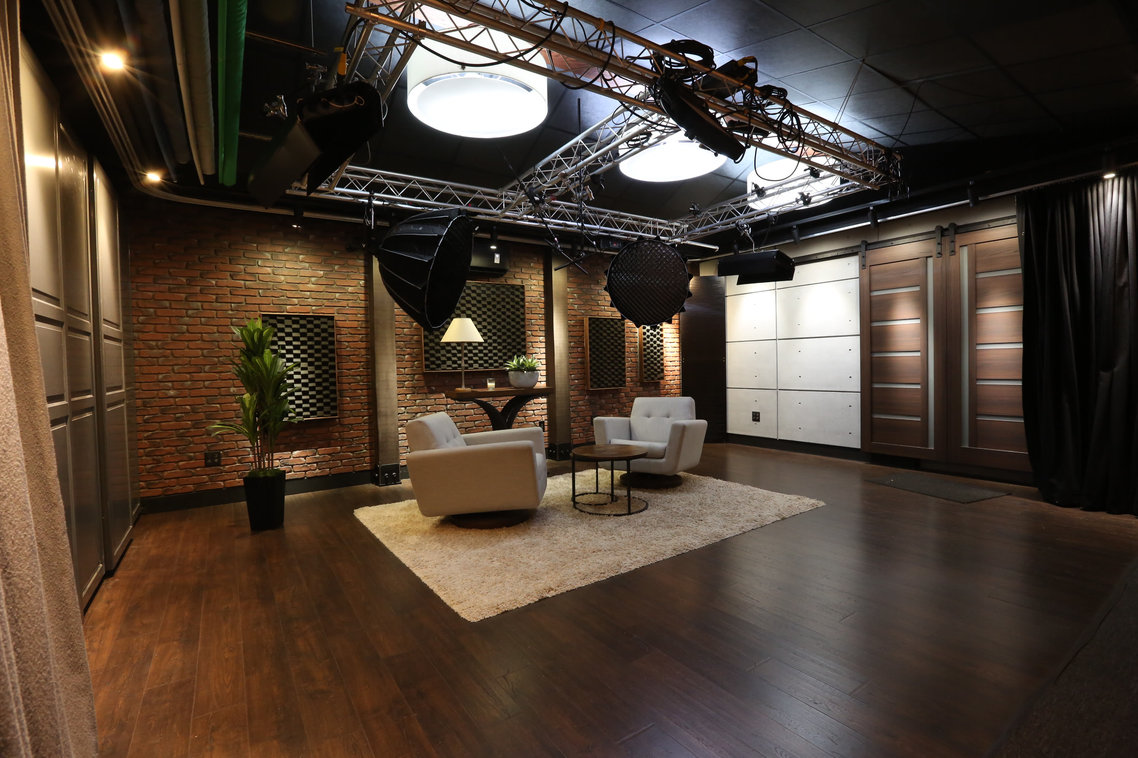 The image shows a modern studio space with two white chairs and a small table centered on a rug, surrounded by brick and paneled walls, and illuminated by overhead lighting.