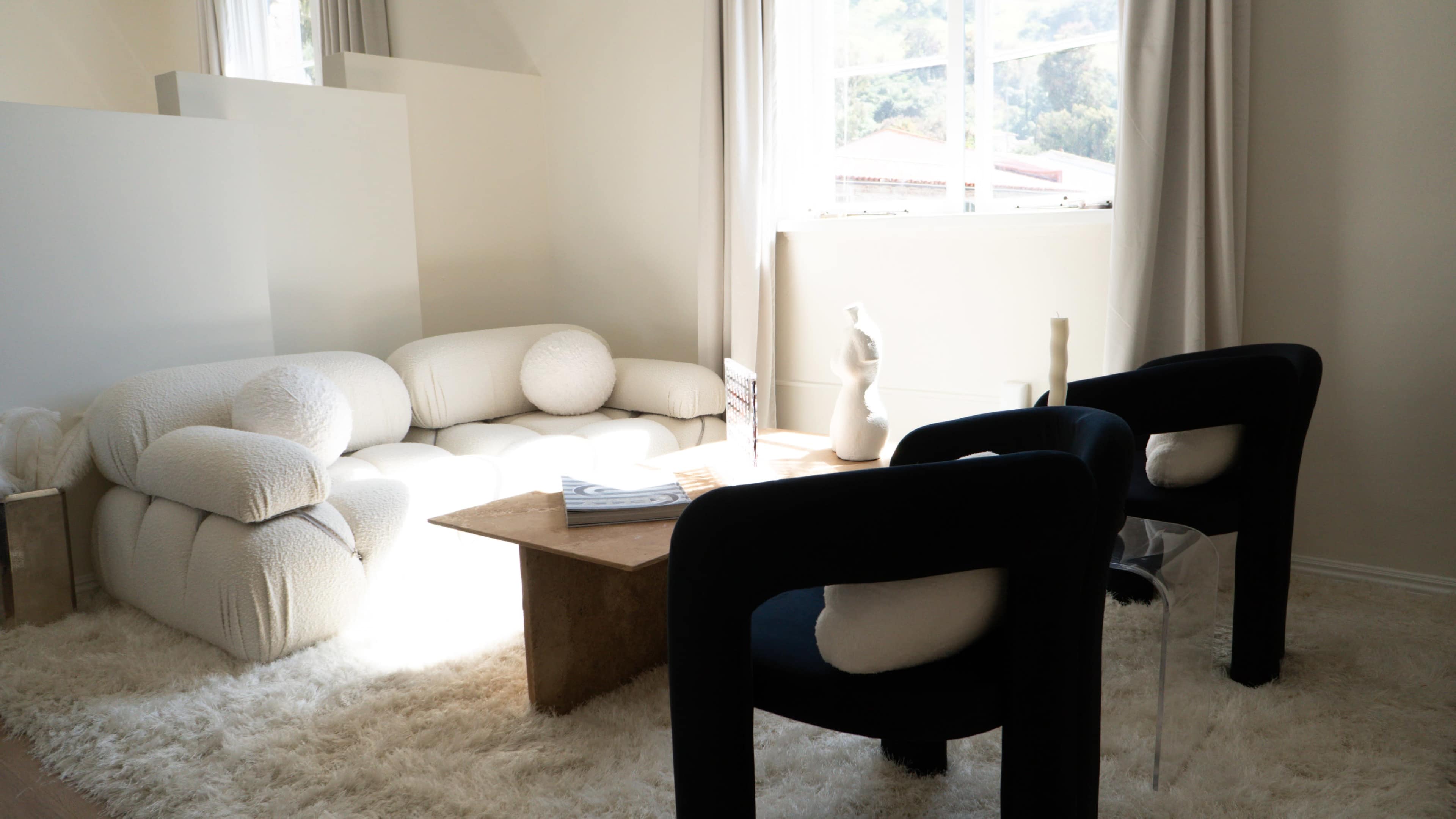 The image depicts a minimalist living room with a cream-colored textured sofa, two black upholstered chairs, a wooden coffee table, and a large window allowing natural light to enter.