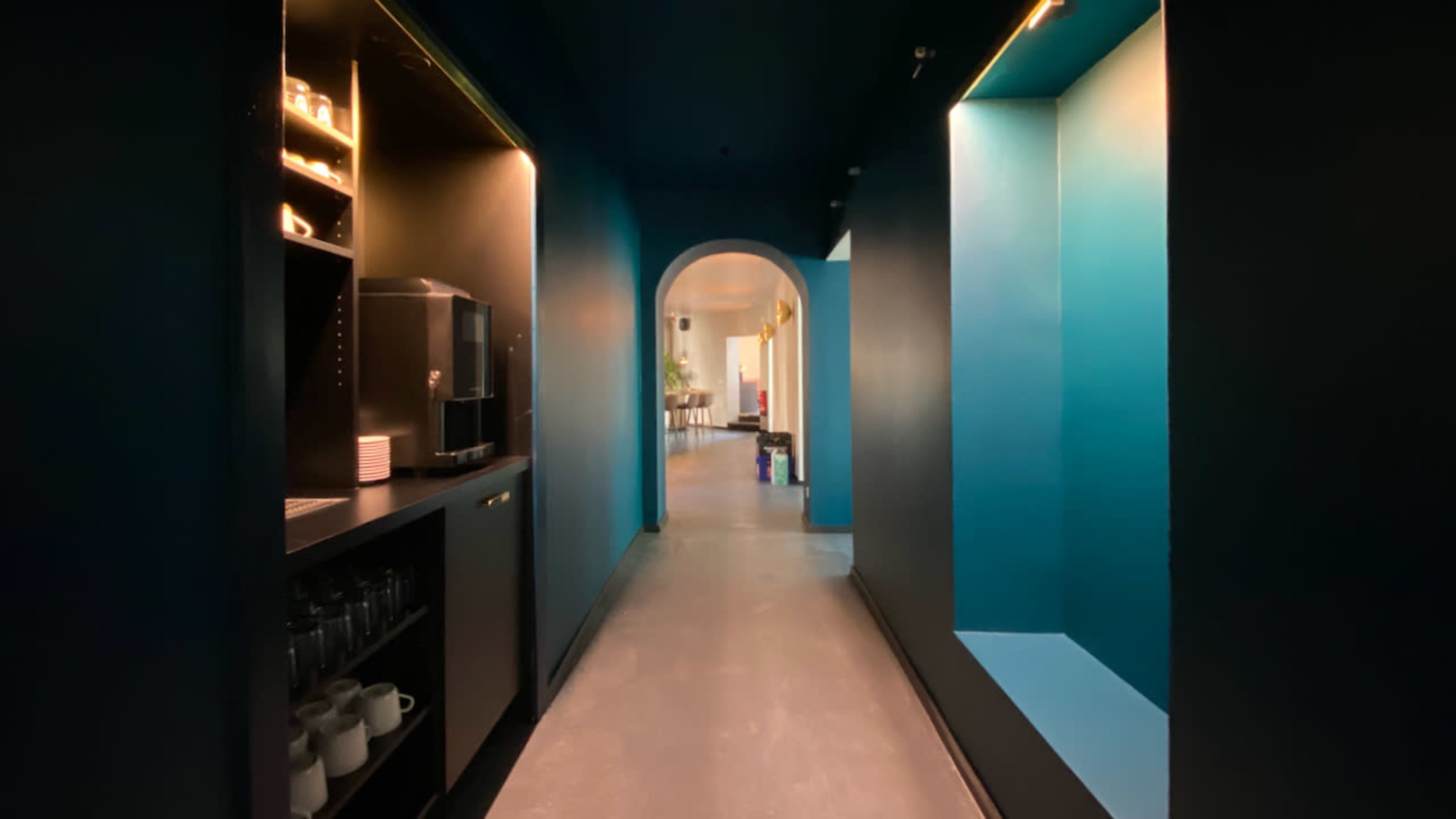 A hallway features blue walls, a coffee station on the left, and illuminated alcoves on the right.