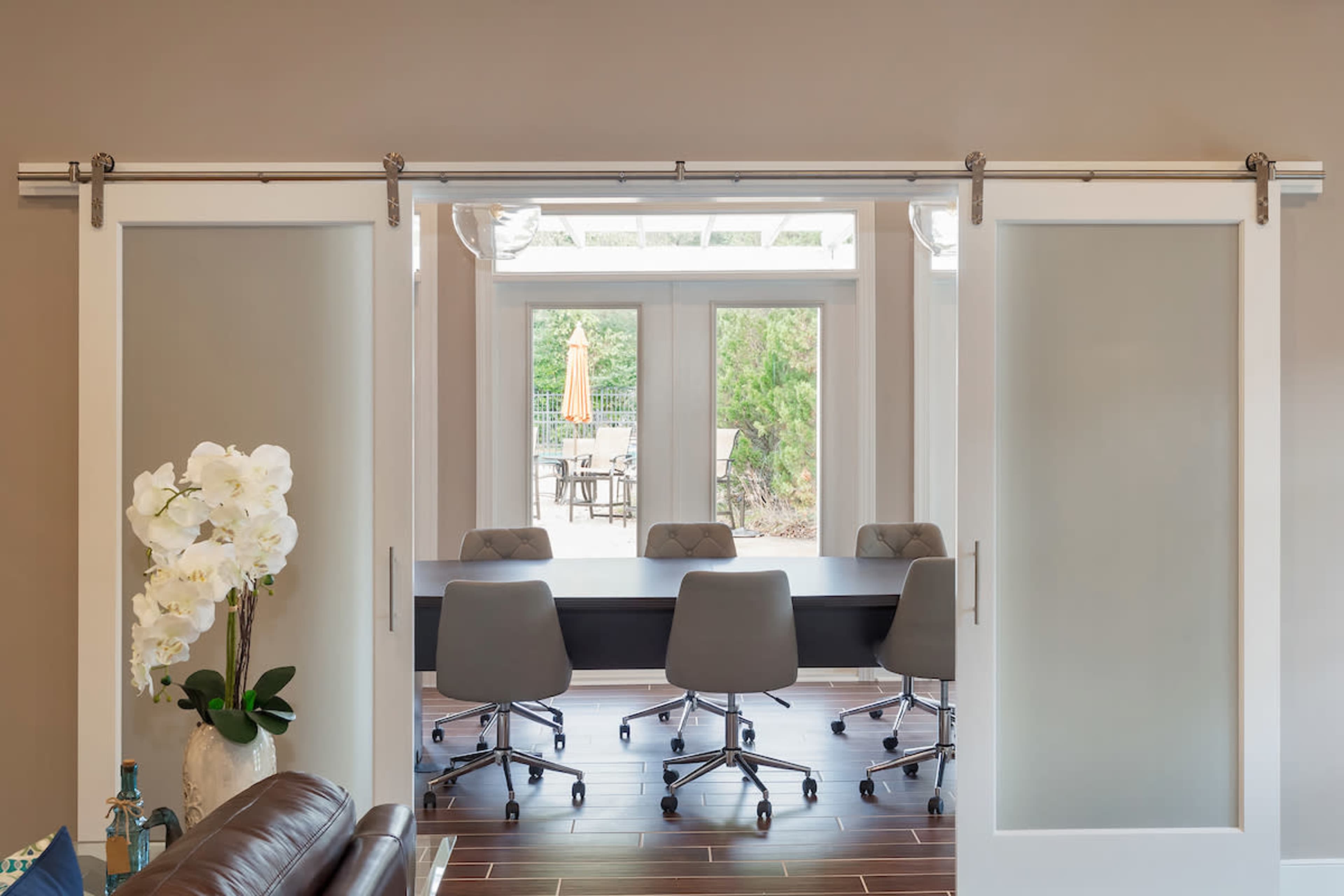 A modern meeting room with a long table and gray chairs, visible through sliding glass doors framed in white.