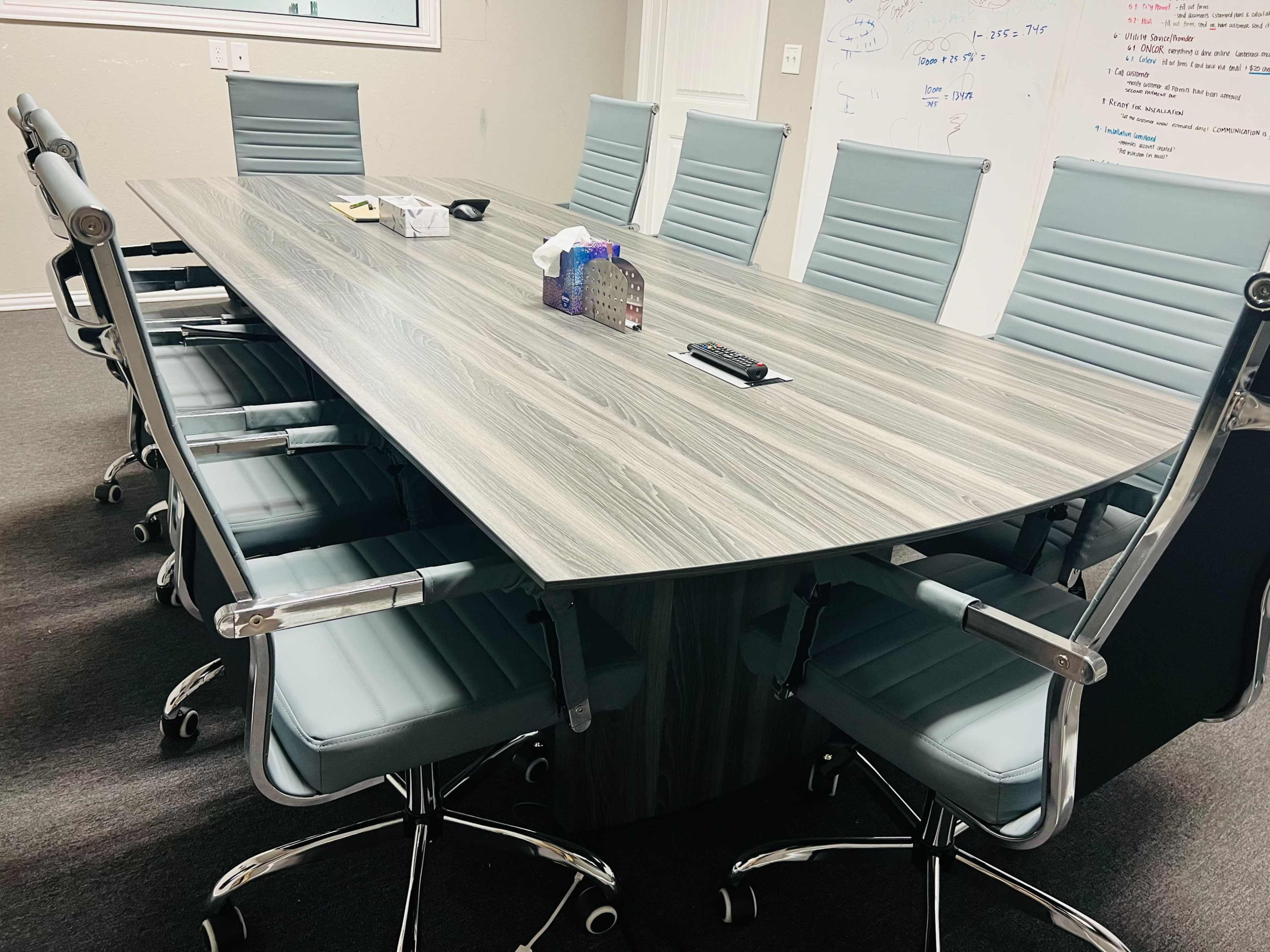 A large conference table is surrounded by modern grey chairs in a meeting room, with a whiteboard visible on one wall.