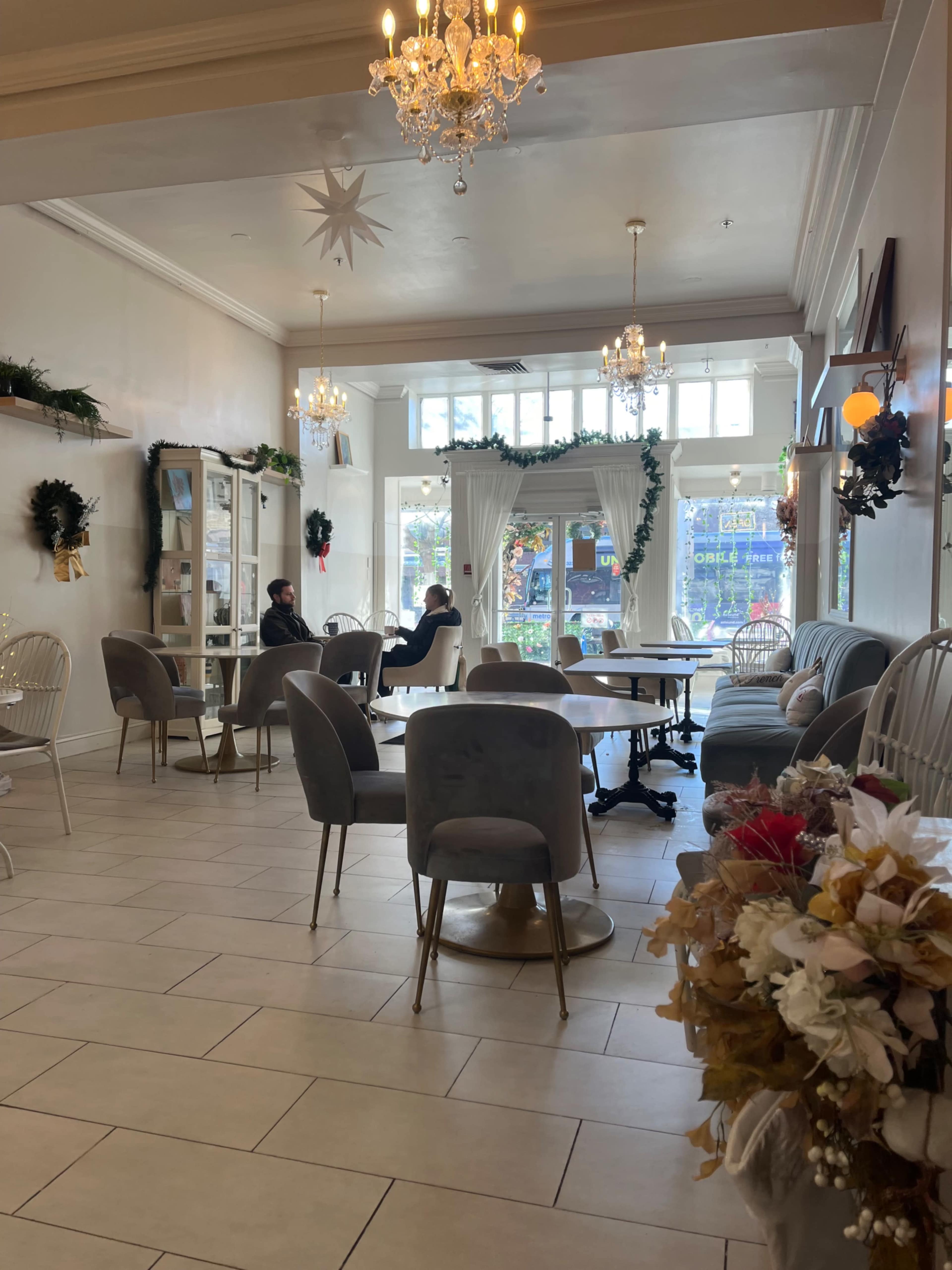 The image shows a cozy café interior with several tables and chairs, large windows letting in natural light, and holiday decorations.