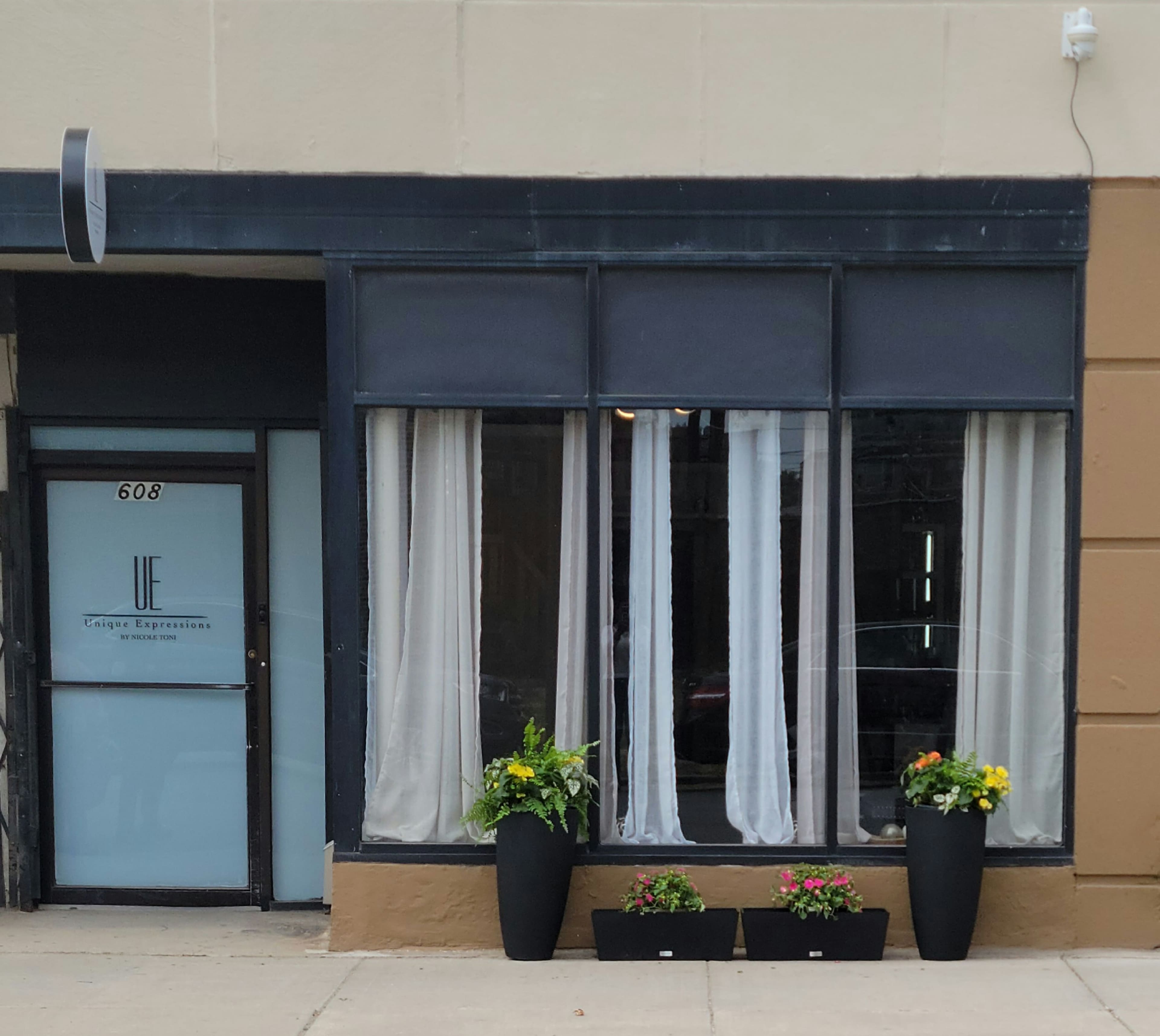 The exterior of a storefront featuring large windows dressed with sheer curtains and two planters with flowers on either side of the entrance.