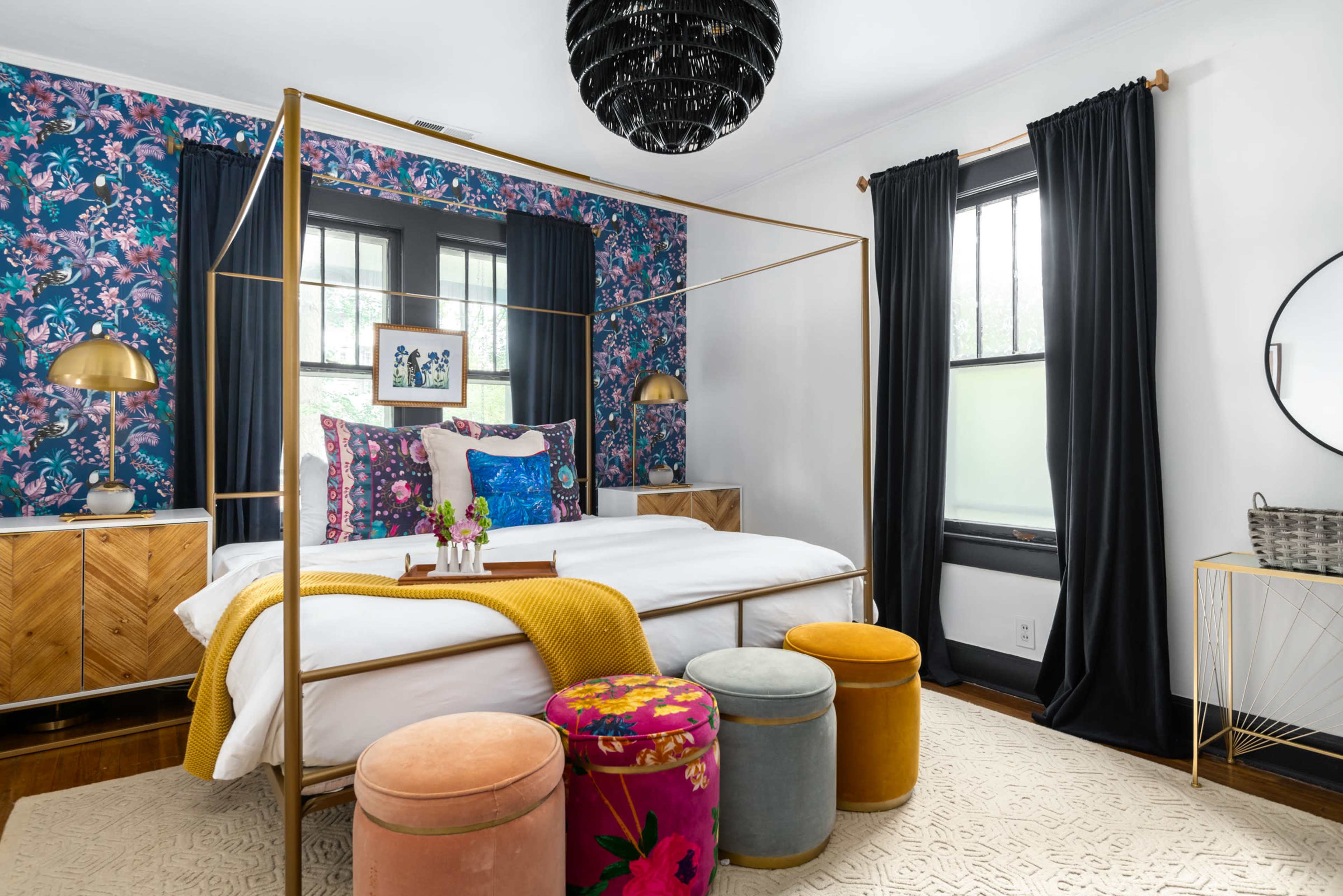 A well-decorated bedroom features a four-poster bed, floral wallpaper, dark curtains, and colorful stools.