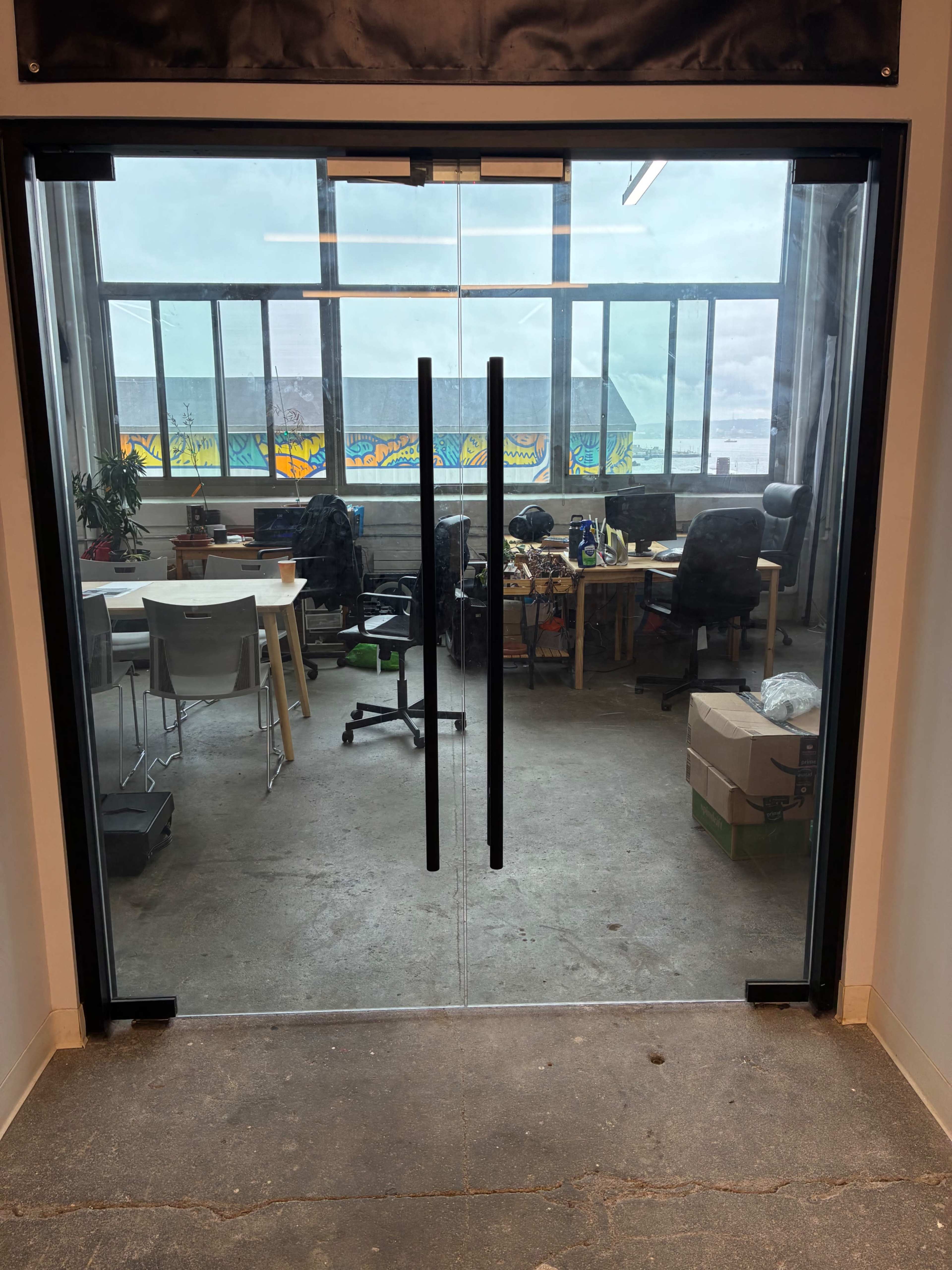 The image shows a pair of glass doors leading to an office space with several desks, chairs, and boxes visible inside.