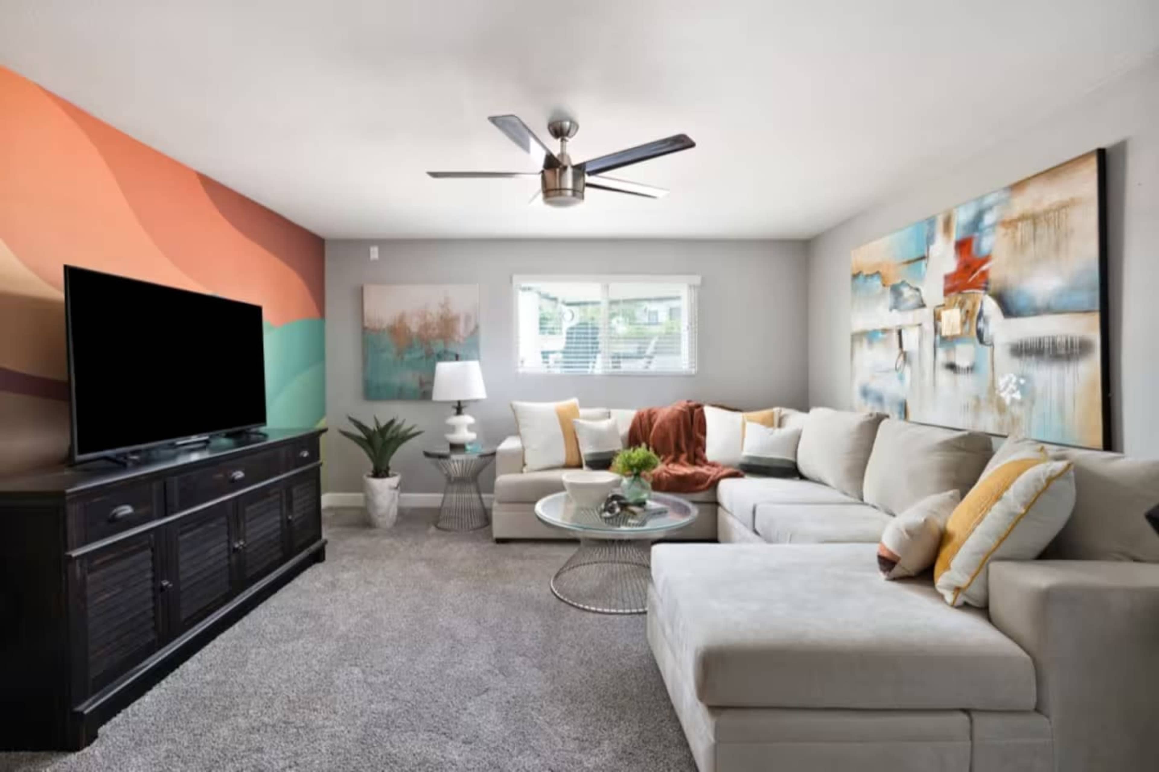 The image shows a modern living room with a beige sectional sofa, a large television on a dark cabinet, and colorful abstract artwork on the walls.