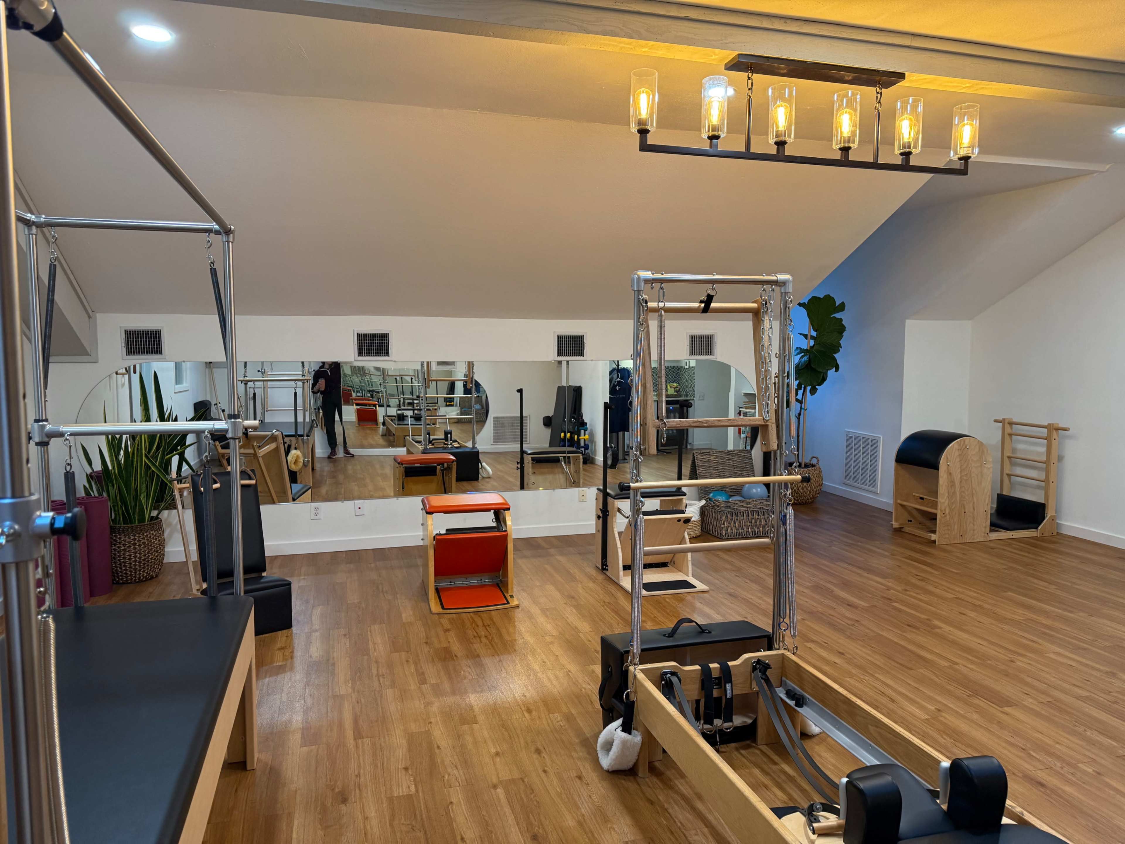 The image shows a well-lit Pilates studio featuring various exercise equipment, mirrors, and a wooden floor.