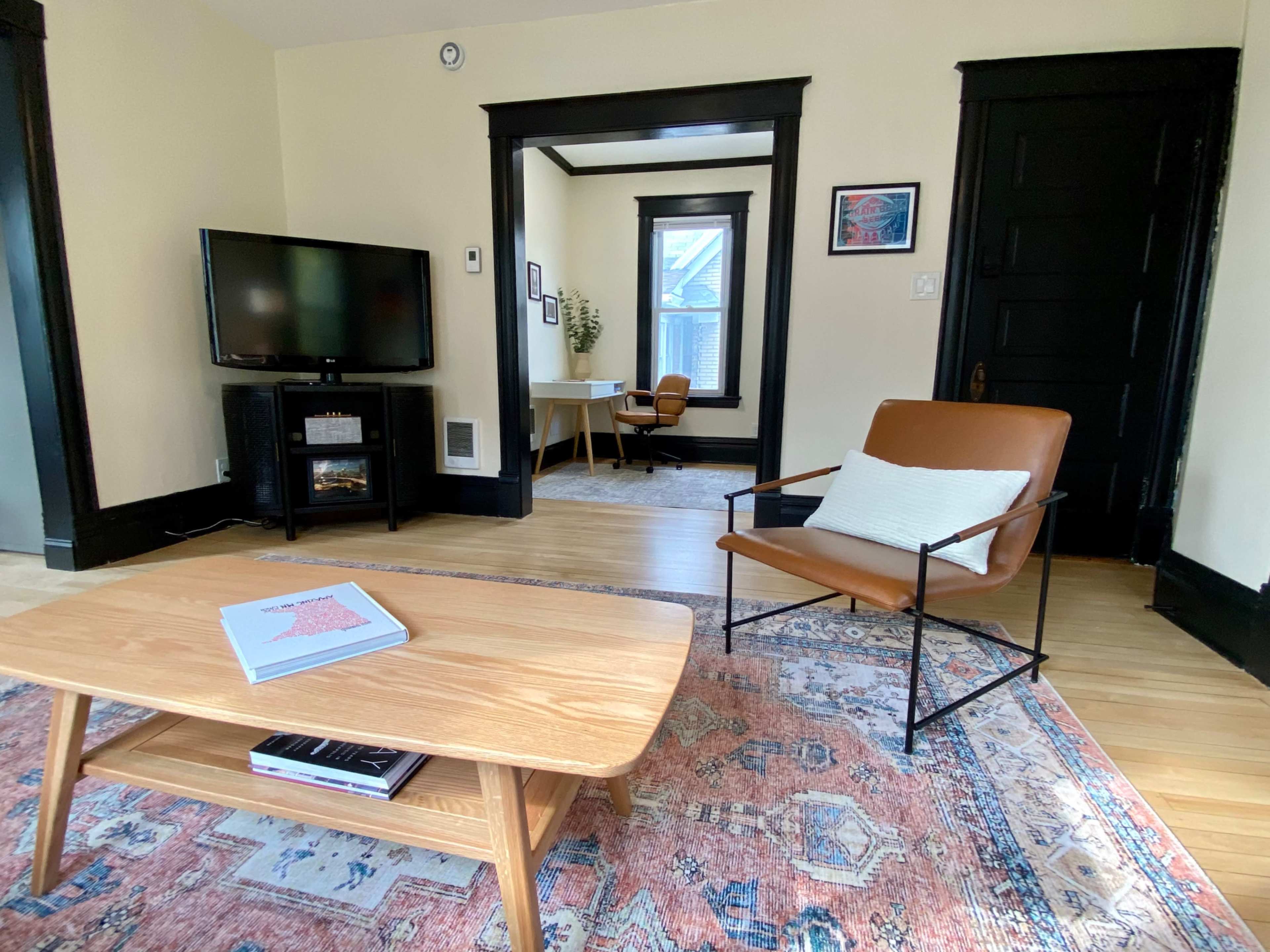 The living room features a wooden coffee table, a leather armchair, a television, and a desk in a bright, organized space.