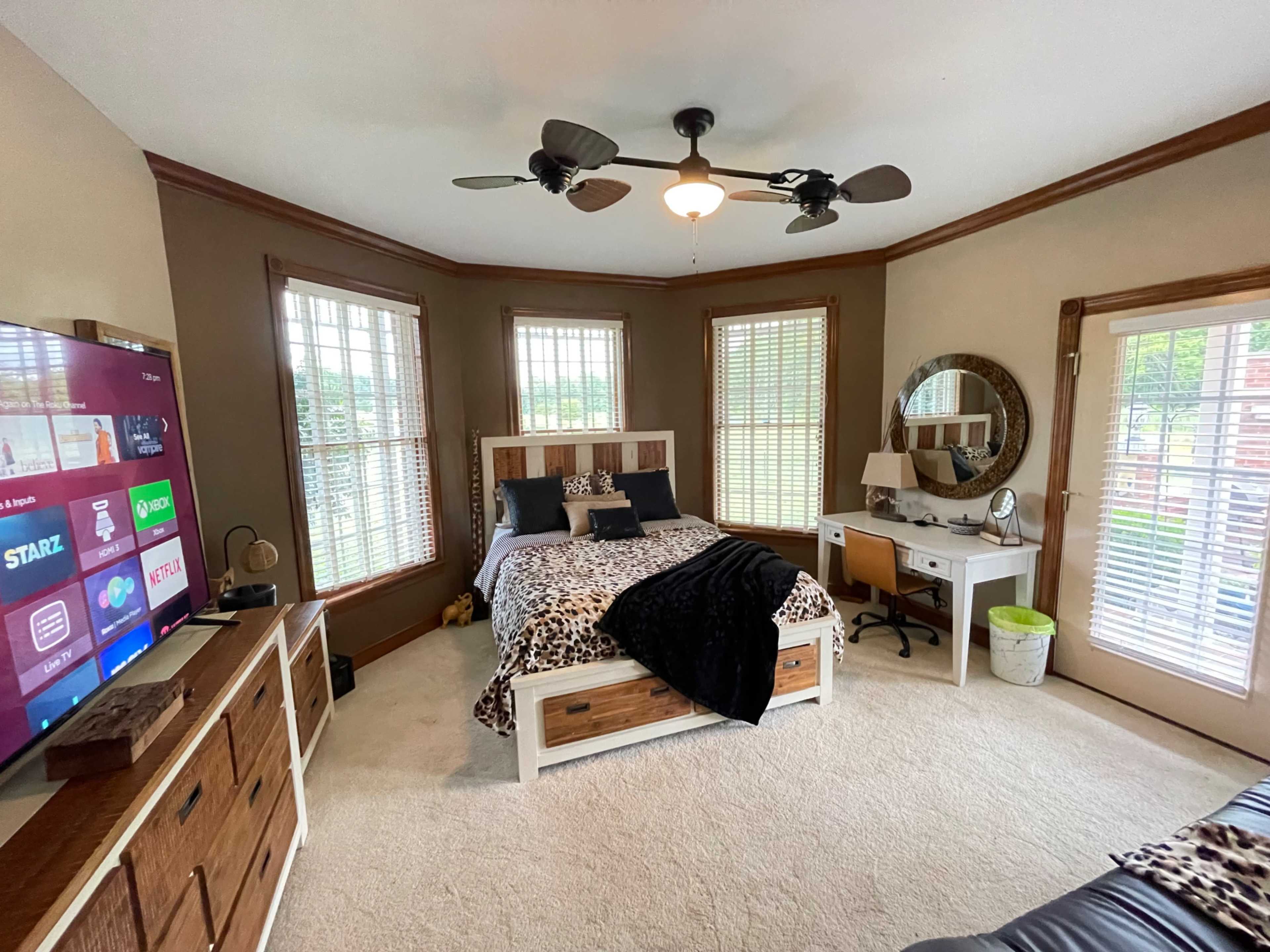 The image shows a bedroom featuring a bed with a leopard print comforter, a wooden dresser, a desk with a lamp, and a large television mounted on the wall.