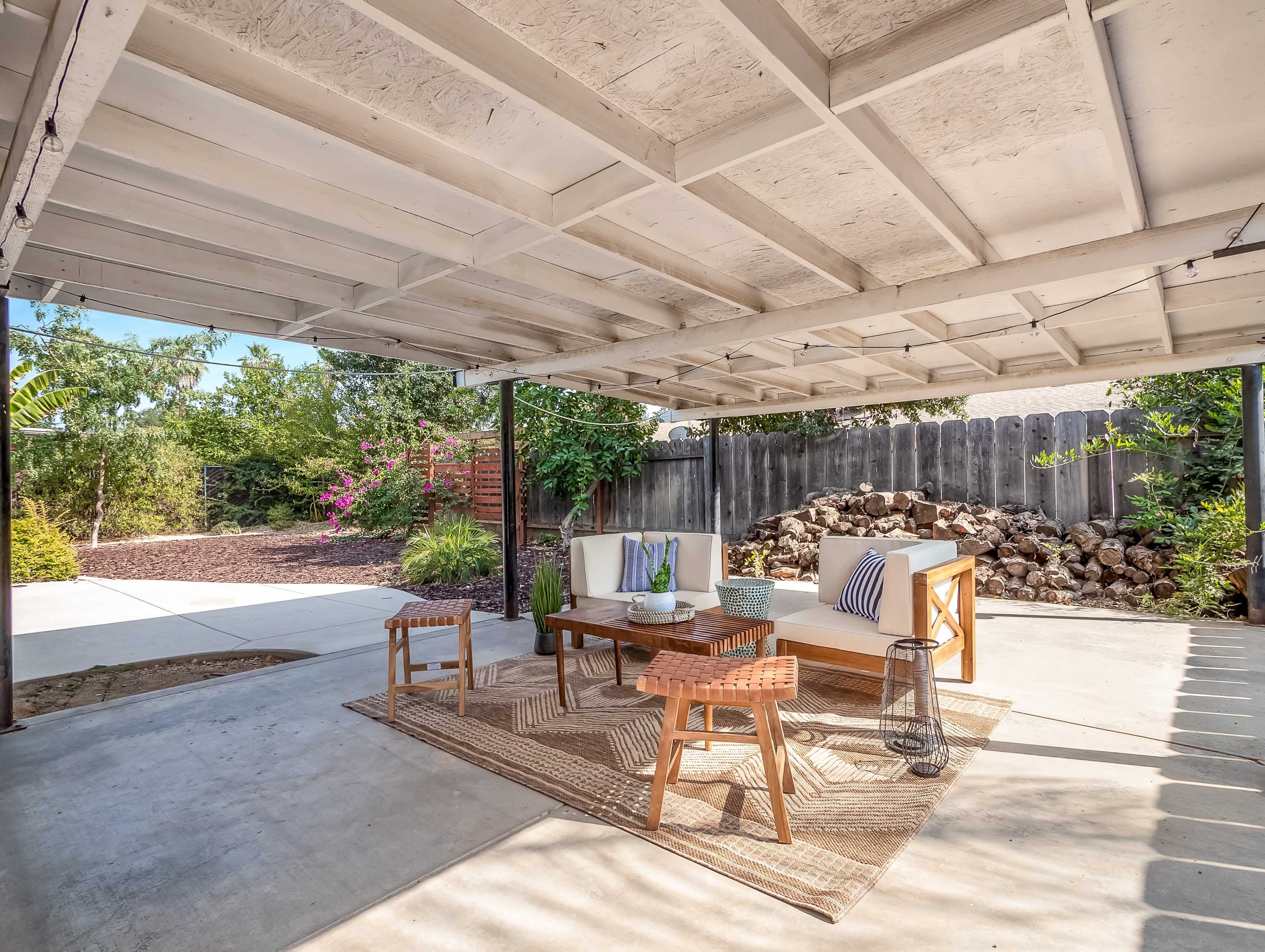 A covered outdoor seating area with a table and chairs surrounded by greenery and a stone wall.