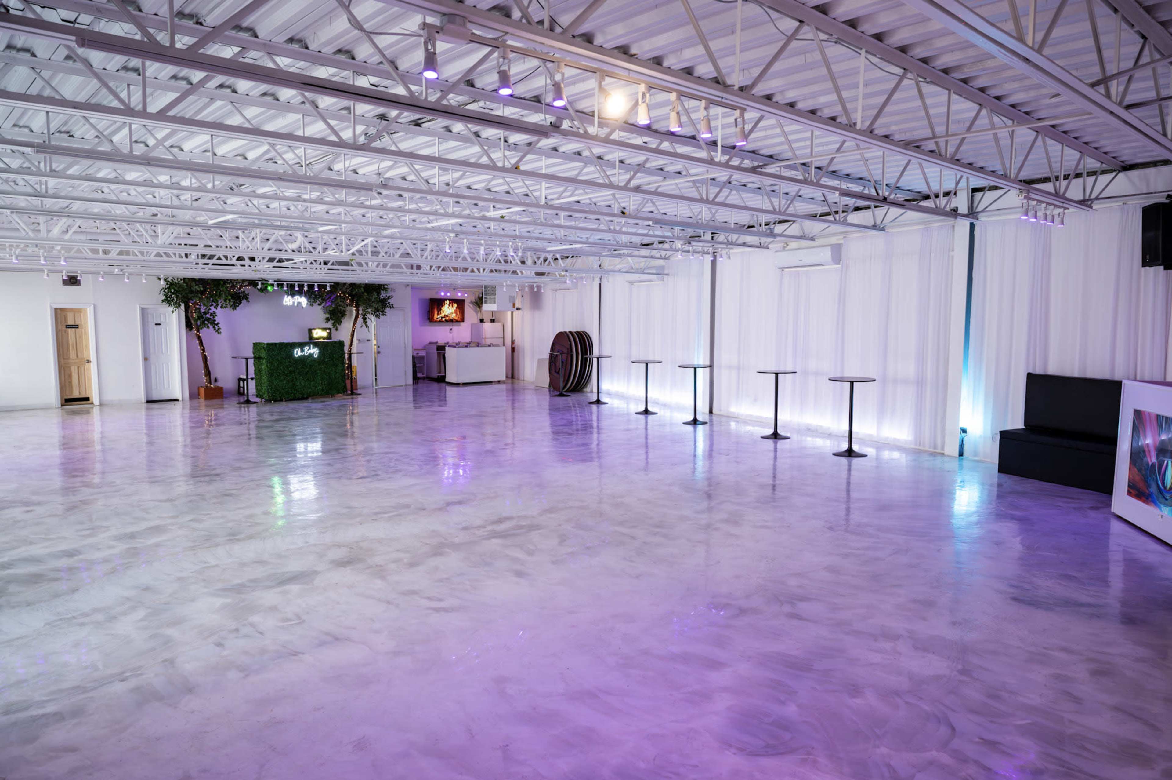 The image shows a spacious, empty event space with polished concrete floors, minimal furnishings, and soft purple lighting.