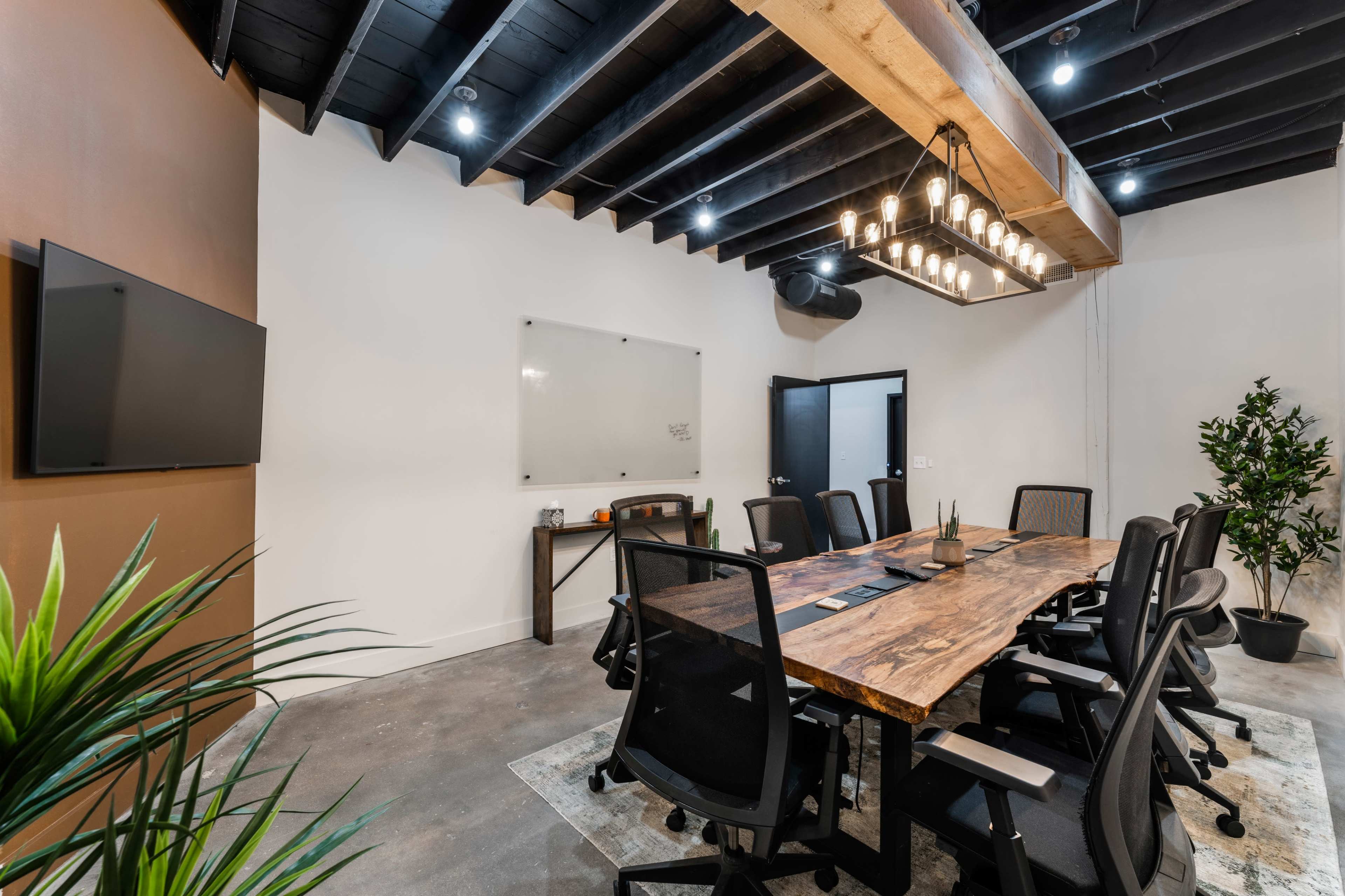 A conference room features a long wooden table surrounded by black chairs, with a whiteboard, two screens, and plants in the decor.