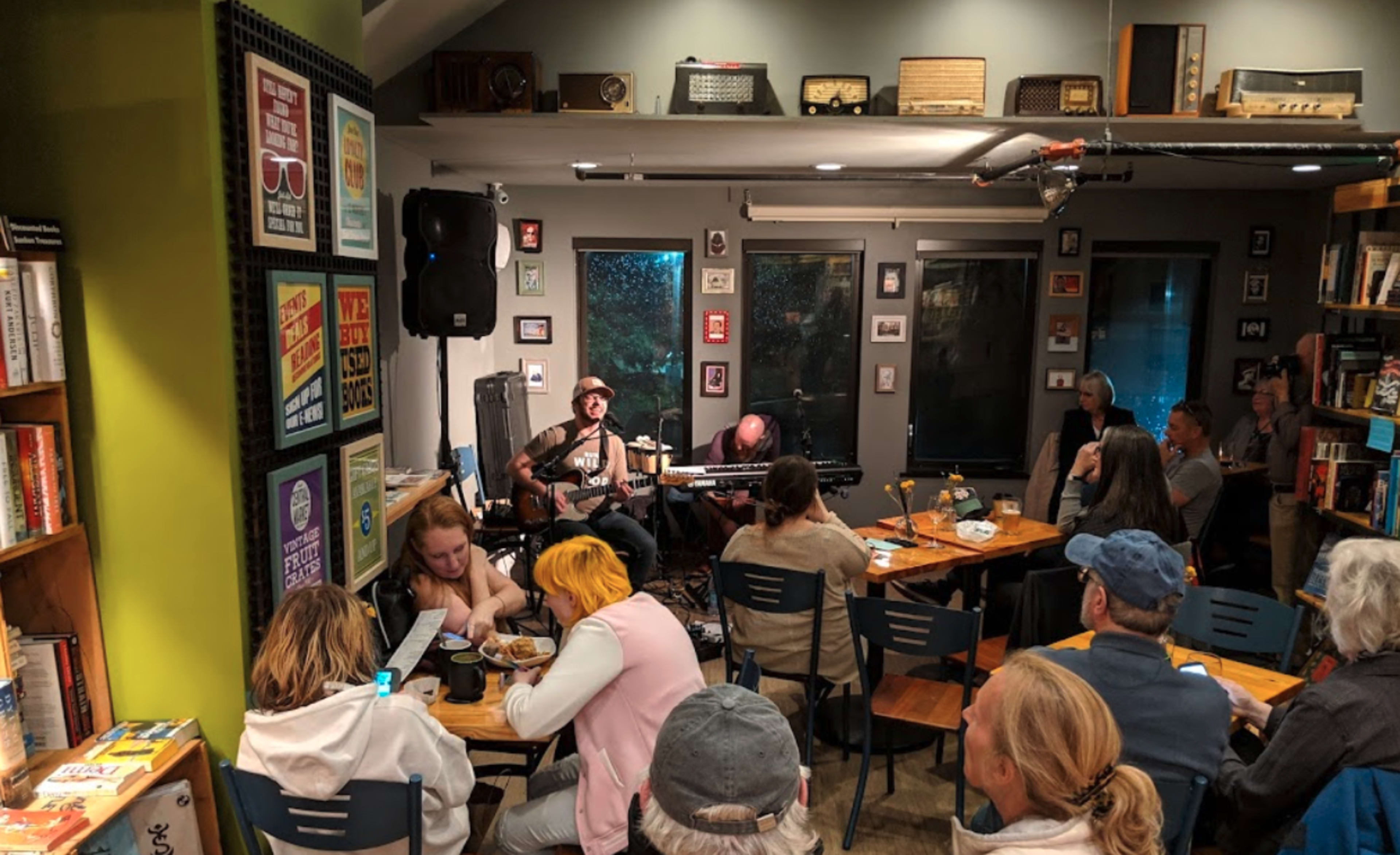 A small crowd listens to a musician performing with a guitar in a cozy cafe decorated with vintage items and framed posters.