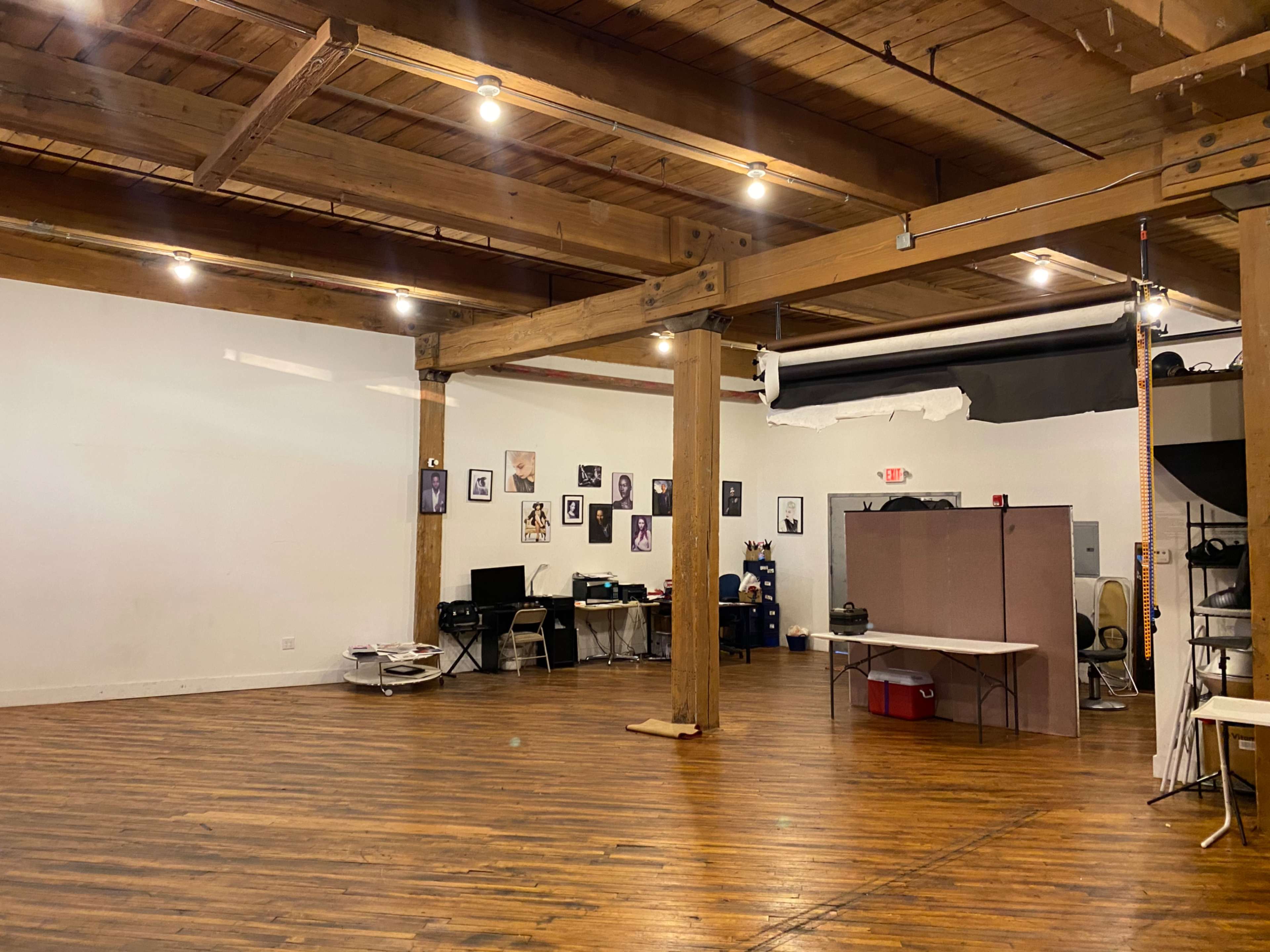 A spacious room with wooden floors and exposed beams, featuring a few desks and a wall adorned with framed photographs.