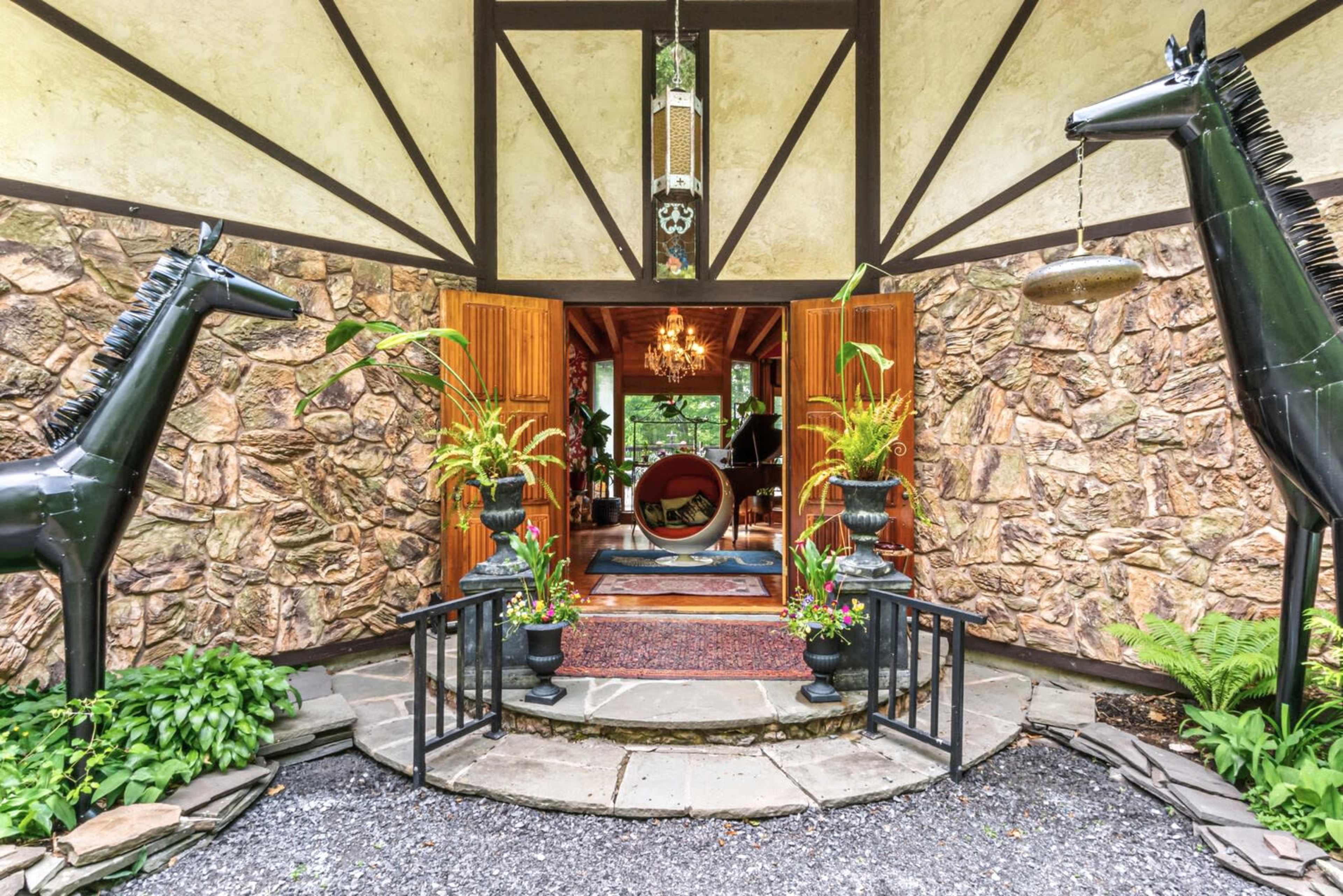 The entrance of a stone house is flanked by two black horse sculptures, leading to a welcoming interior with a round seating area and decorative plants.