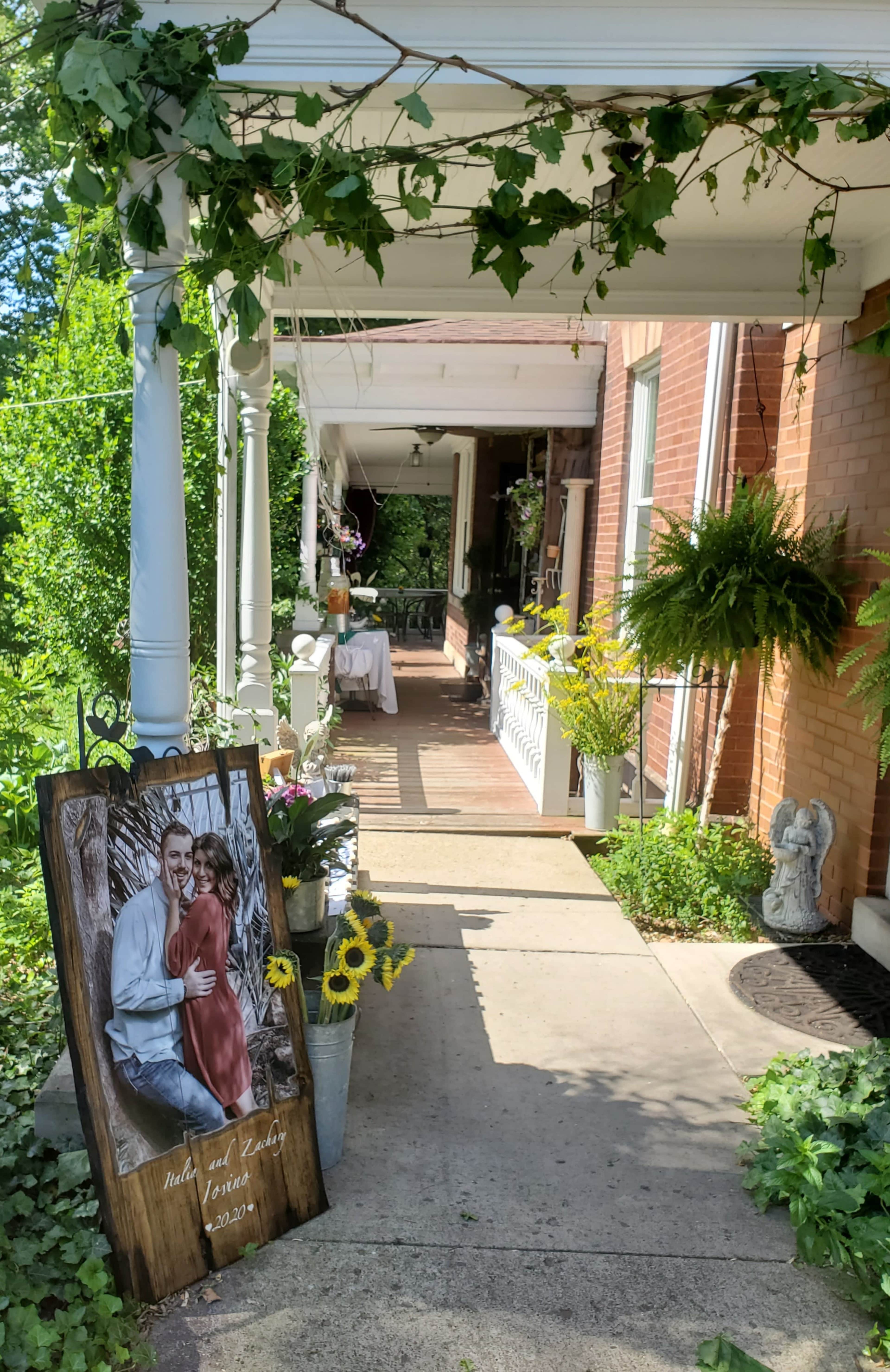 A pathway lined with greenery leads to a porch adorned with a decorative sign and potted plants.