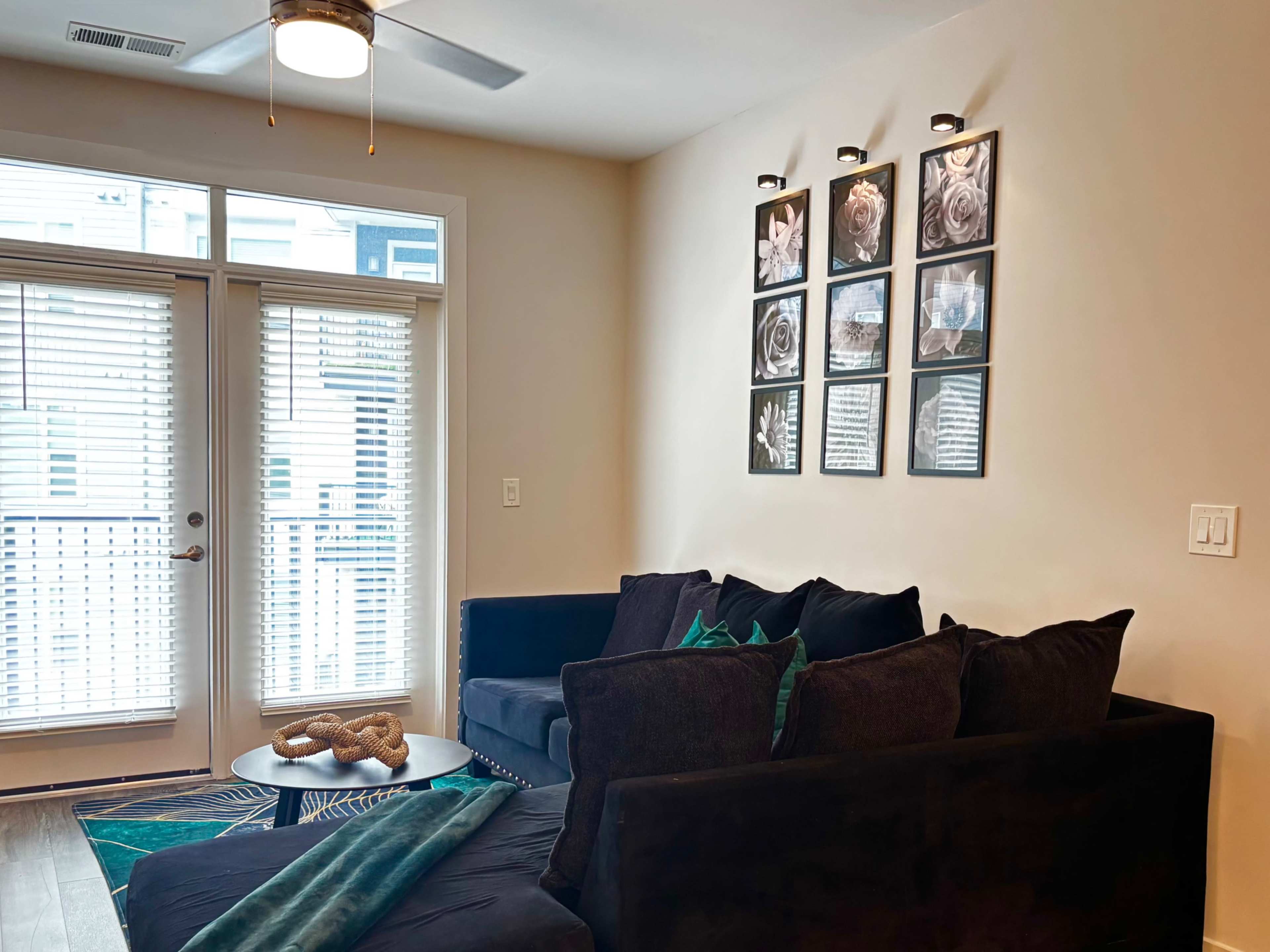 The image shows a living room with a dark sofa, decorative pillows, a coffee table, and framed pictures on the wall, with sliding glass doors leading to a patio.