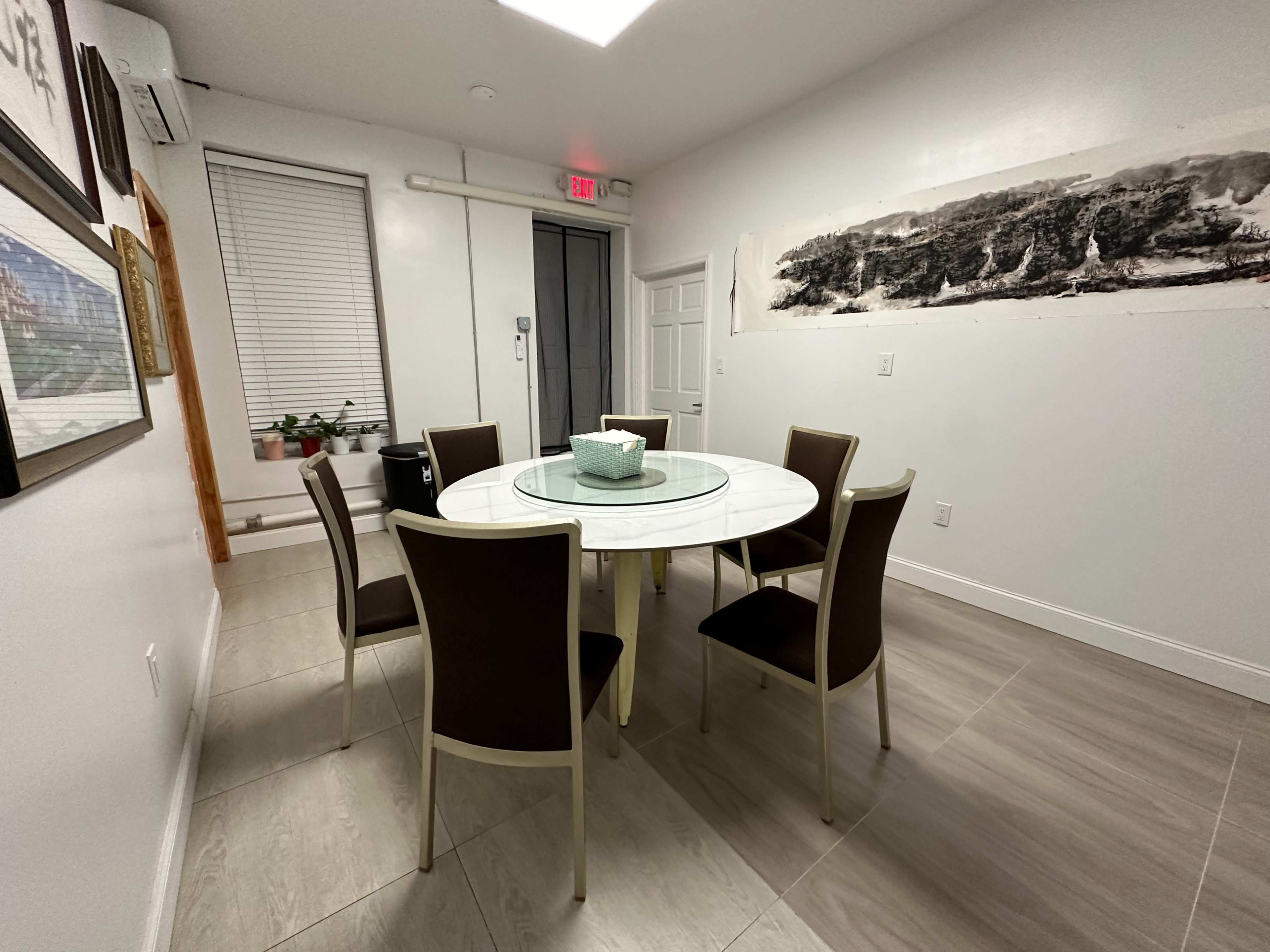 A round glass table with six chairs is seen in a well-lit, minimalist dining area featuring a large wall artwork and a window.