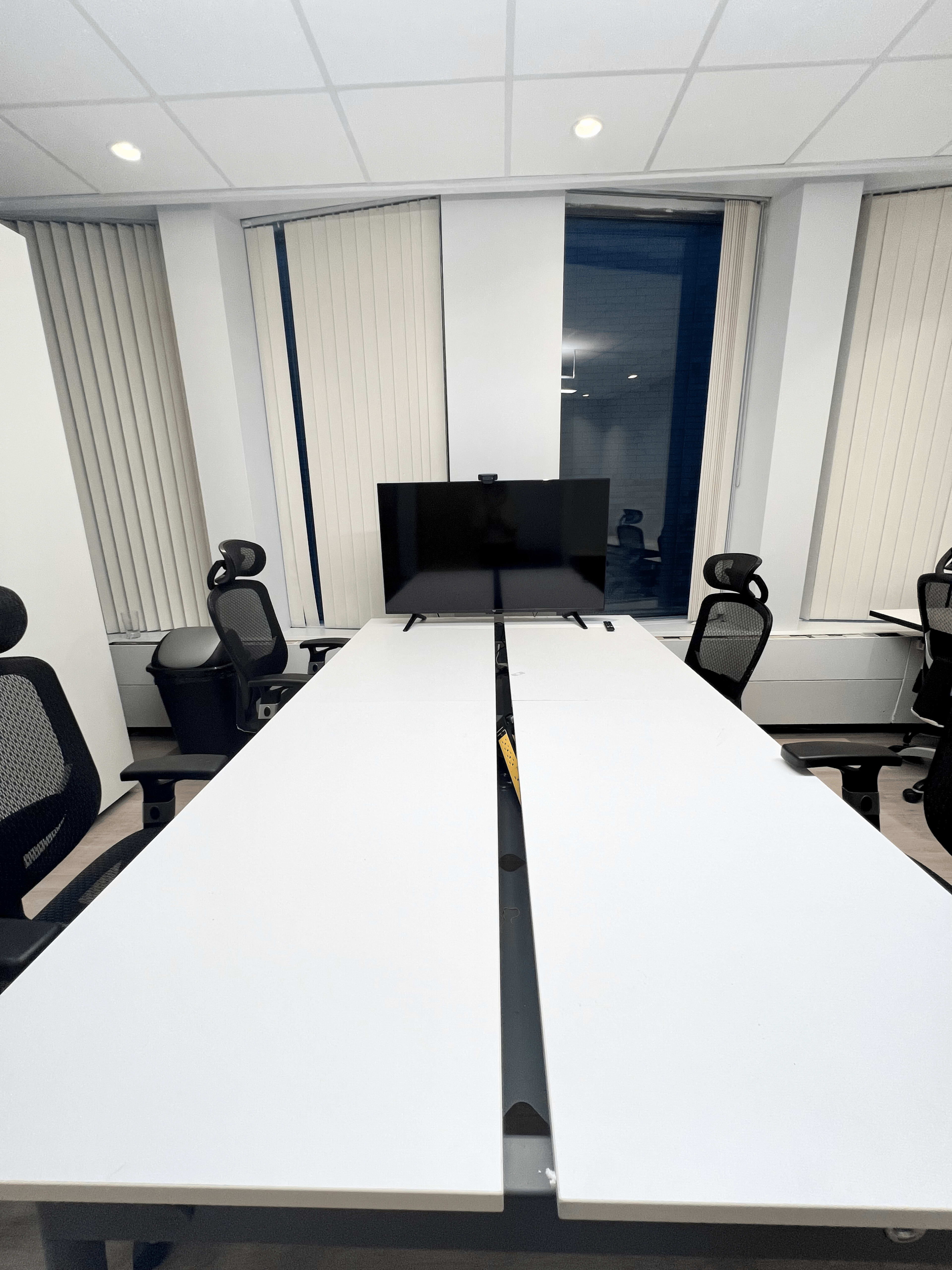 A long conference table is positioned in a brightly lit room with office chairs and a large monitor at one end.