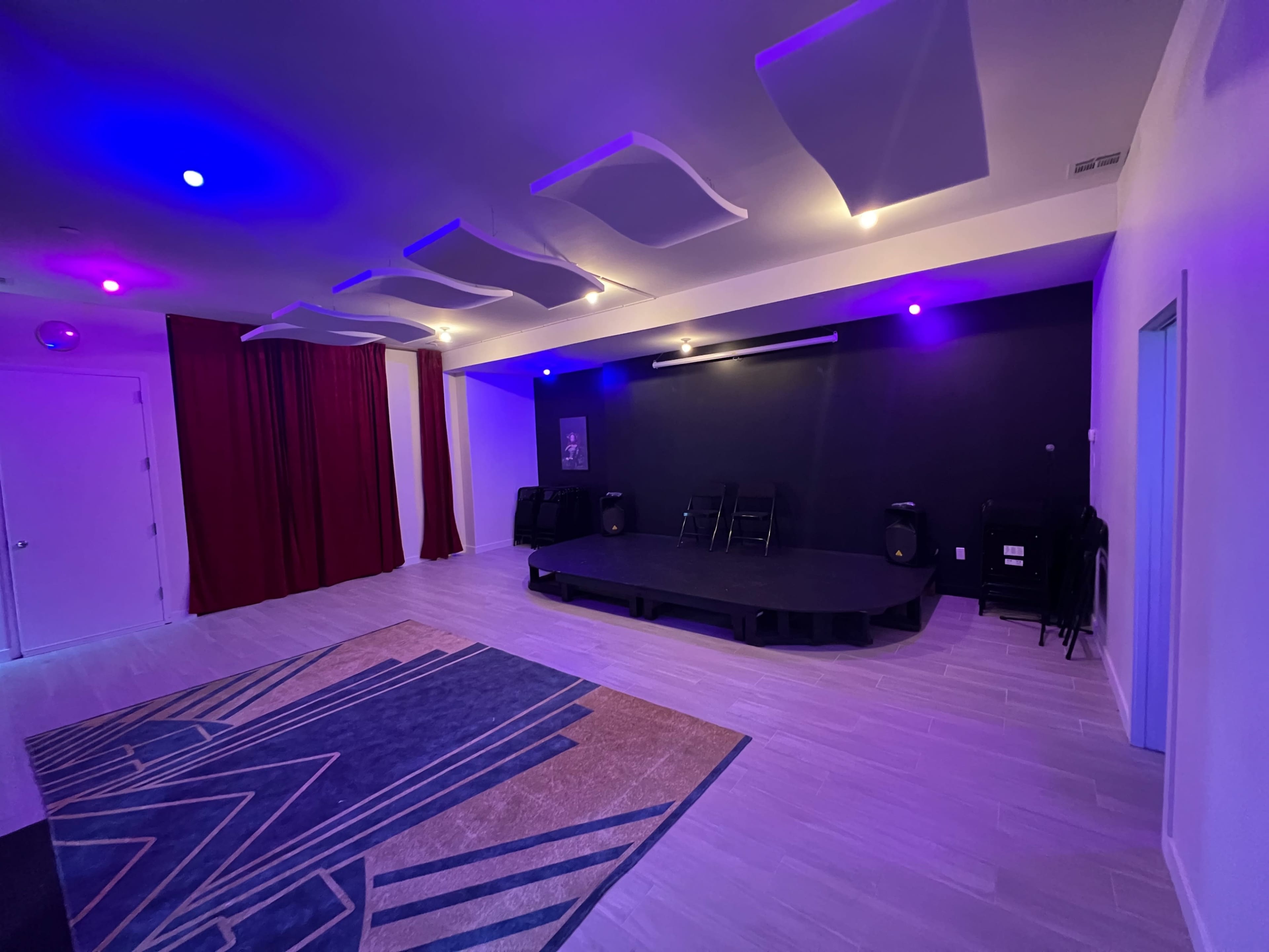 A spacious room with a stage, purple lighting, and sound equipment, featuring a red curtain on one side and a patterned carpet on the floor.