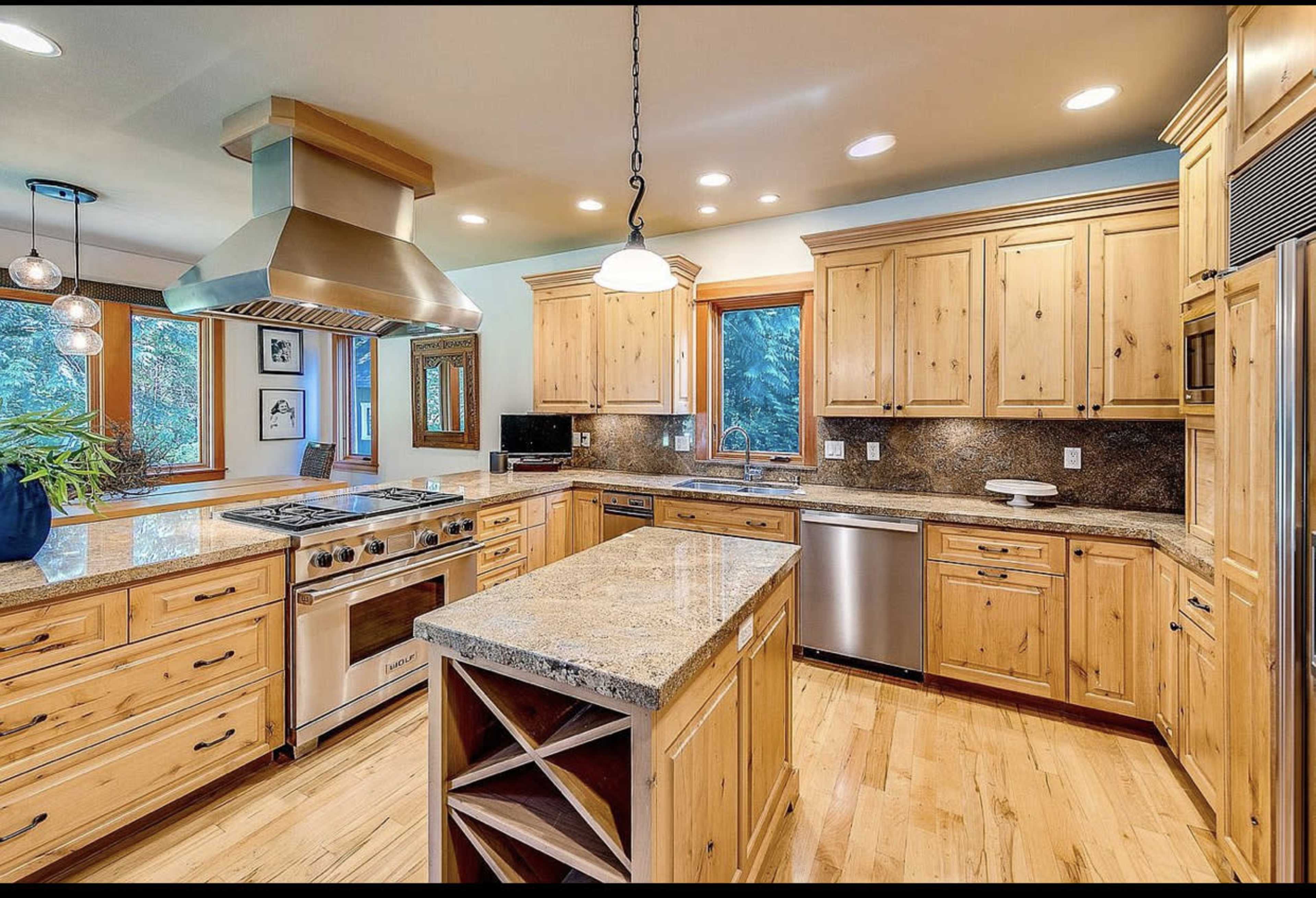The kitchen features wooden cabinetry, a large island with a wine rack, and stainless steel appliances beneath a hooded range.