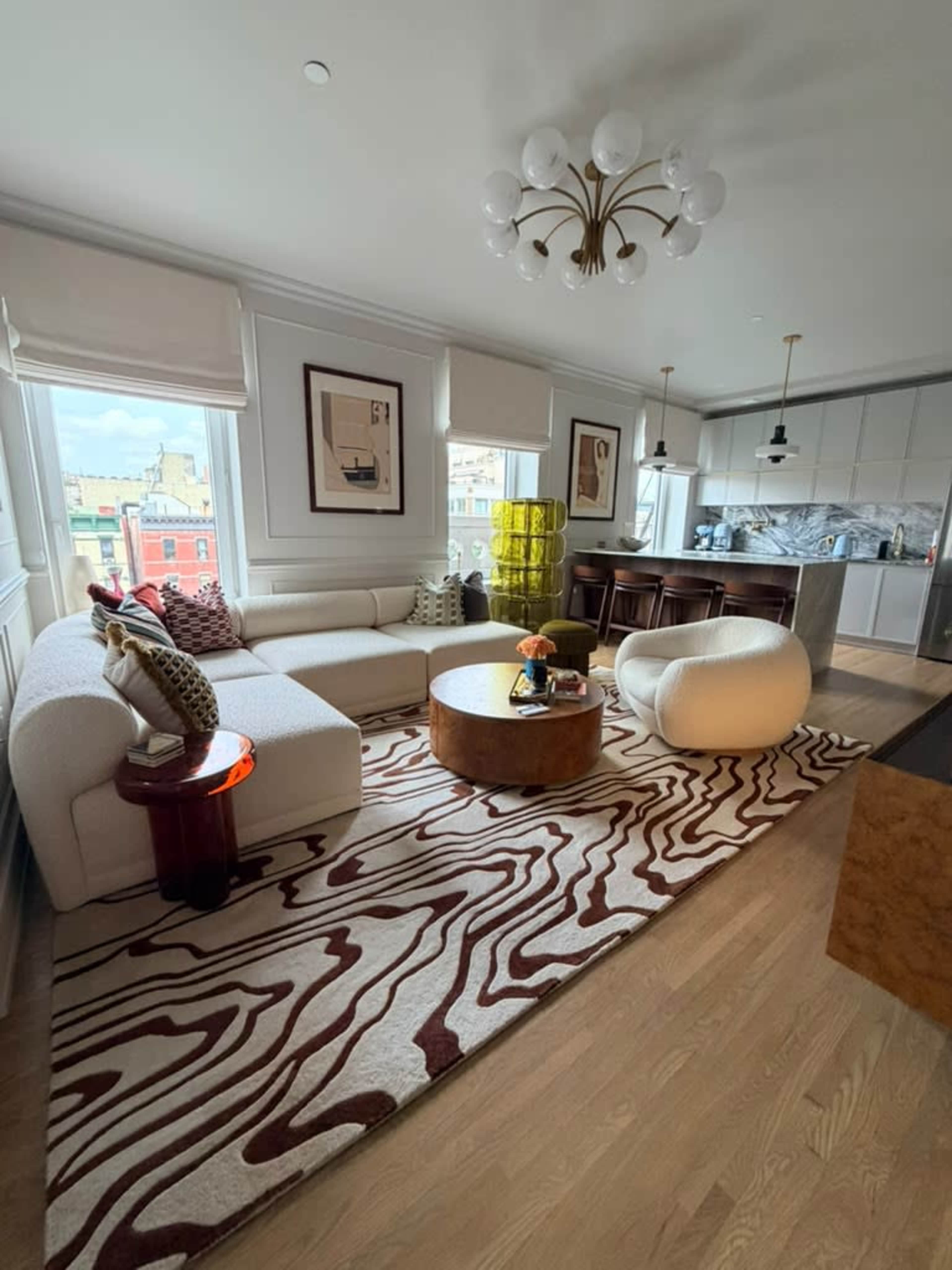 The image shows a modern living room with a sectional sofa, round coffee table, and a large rug, featuring large windows that provide natural light and a view of the outside.