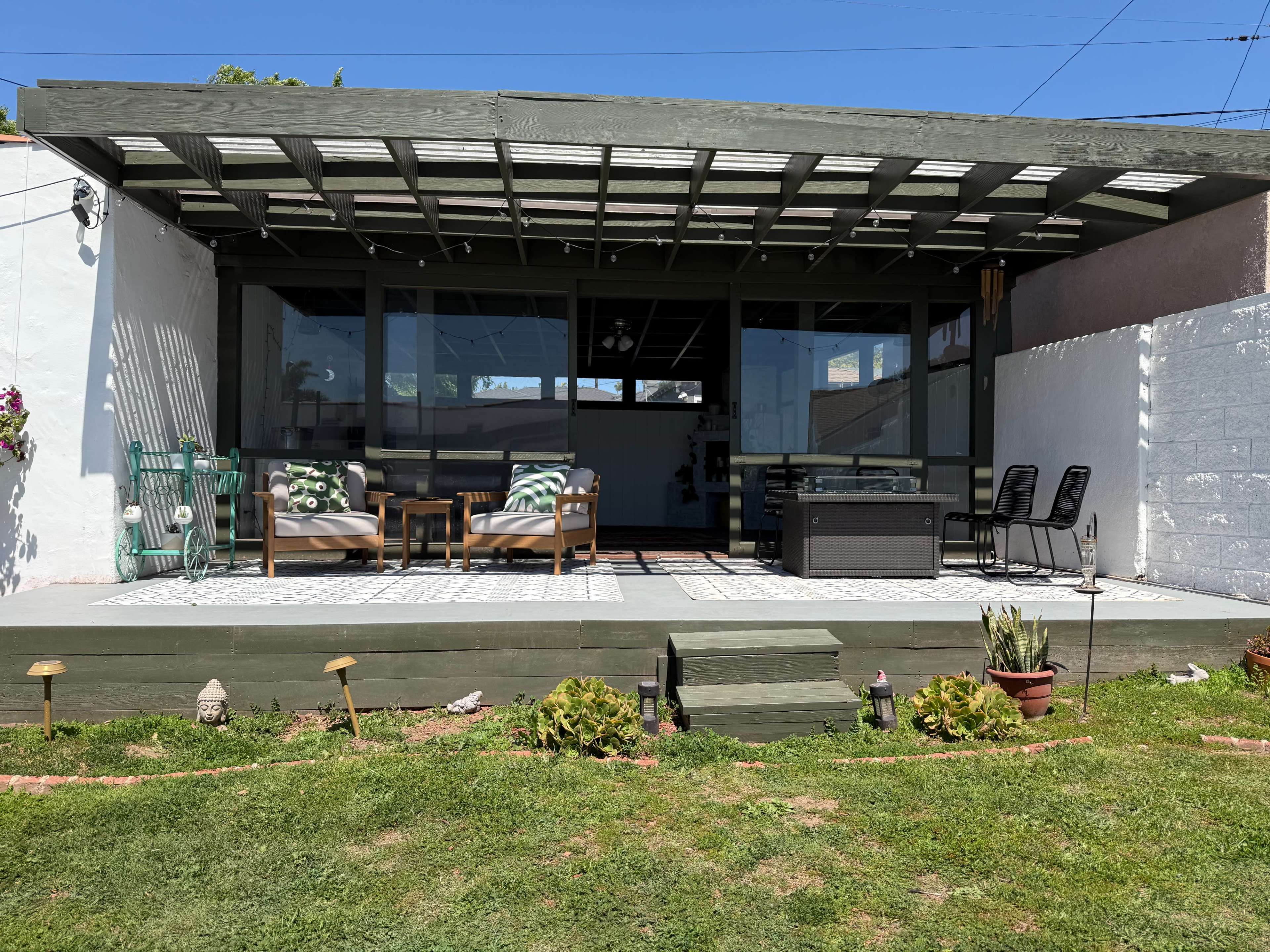 The image shows a home patio with a covered sitting area featuring two chairs, a table, and a decorative floor, surrounded by greenery and a white wall.