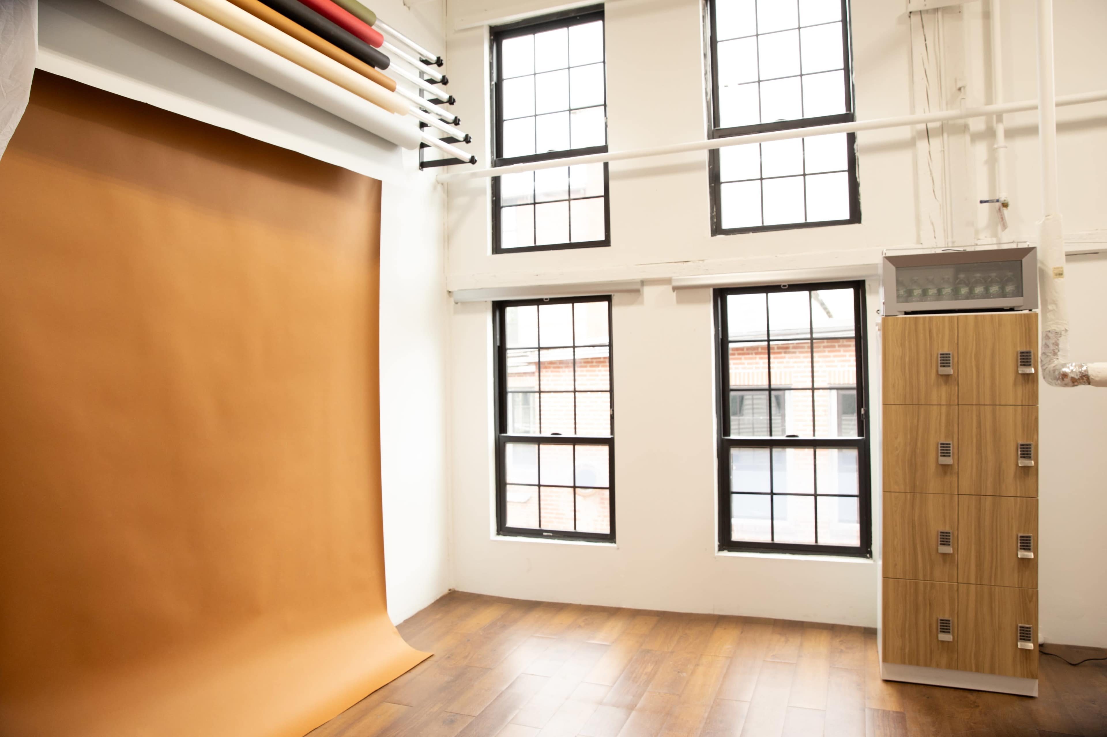 The image shows a photography studio with a brown backdrop draped on the floor and large windows allowing natural light to enter.