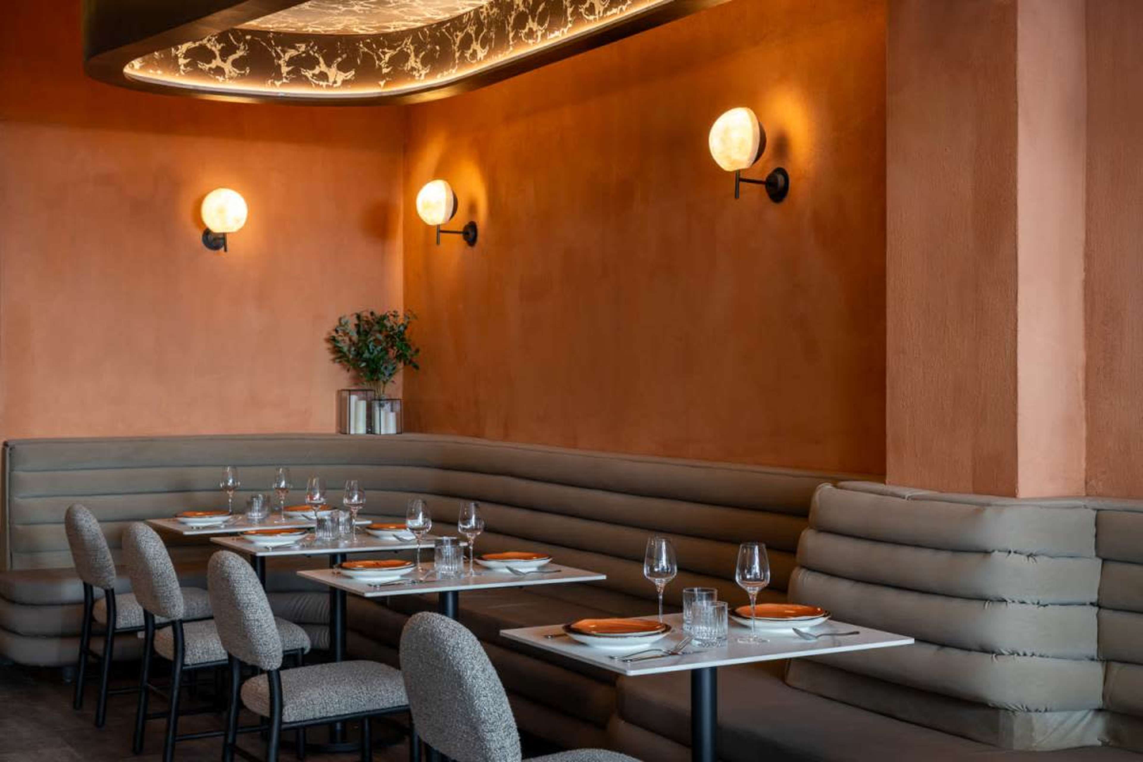 The image shows a stylish restaurant interior featuring orange walls, modern lighting fixtures, and neatly arranged tables with seating.