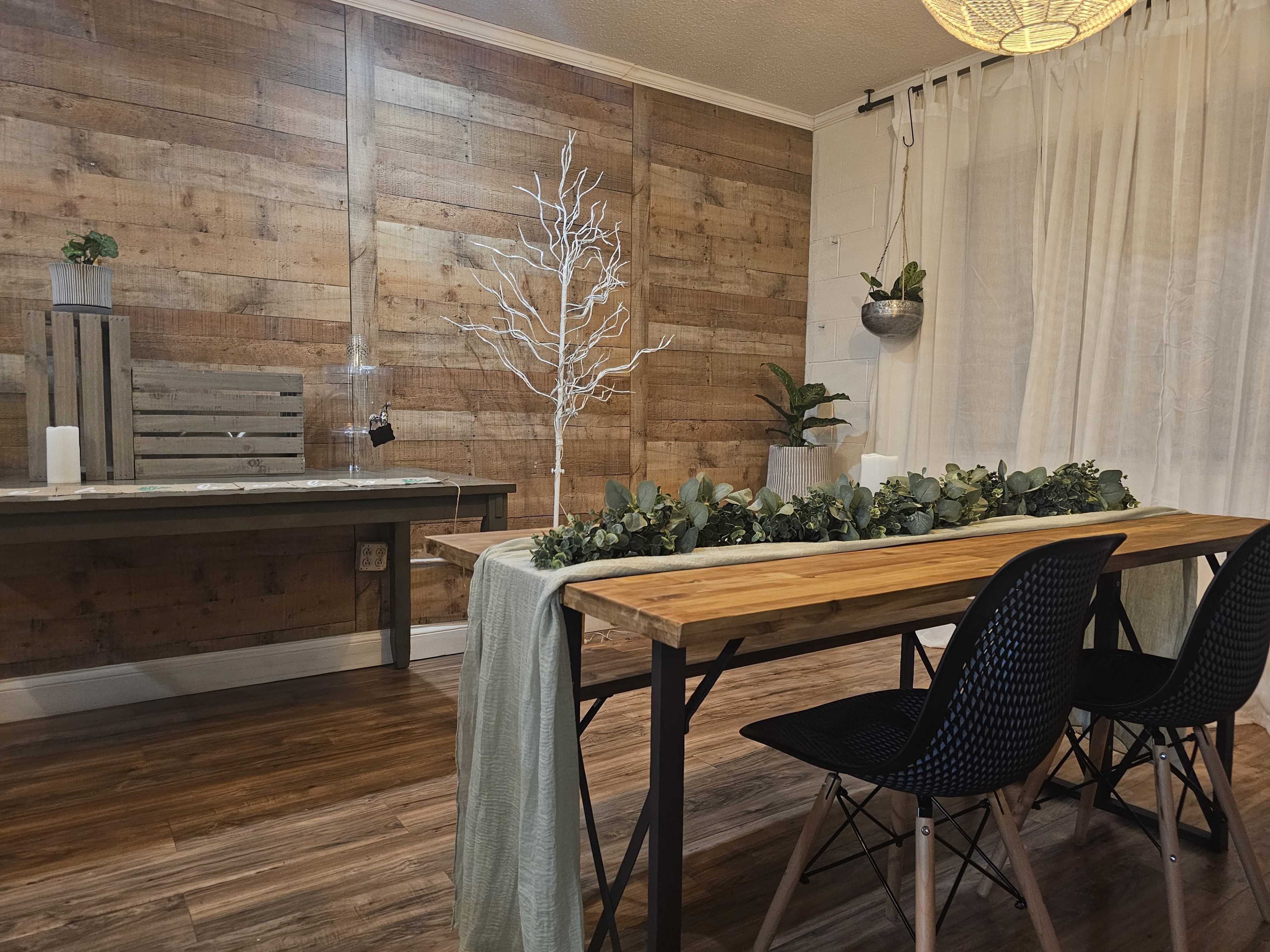 A rustic dining area with a wooden table adorned with greenery, black chairs, and a backdrop of a wooden wall and plants.