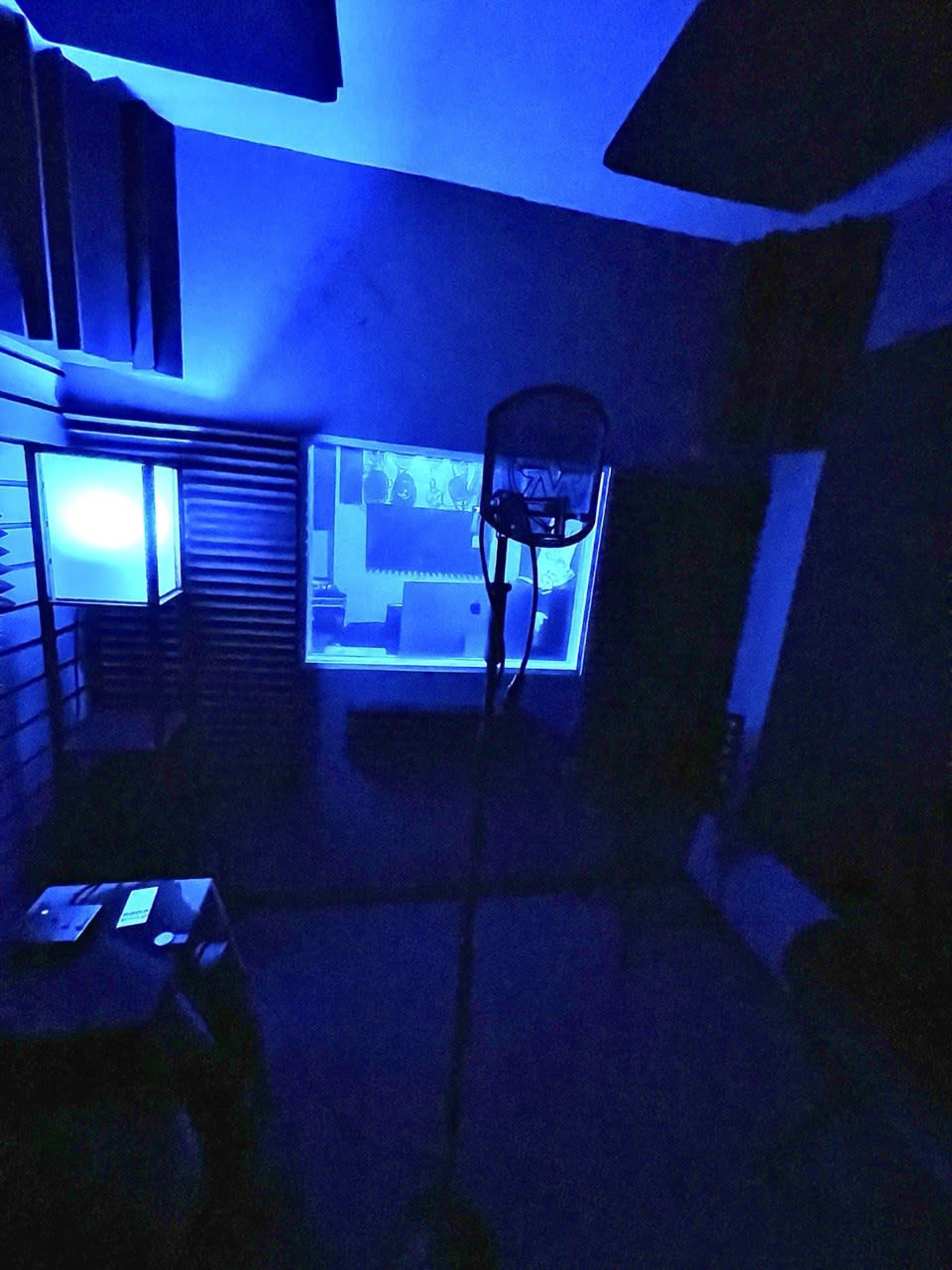 A dimly lit recording studio features a microphone in the foreground and a blue-lit window in the background.