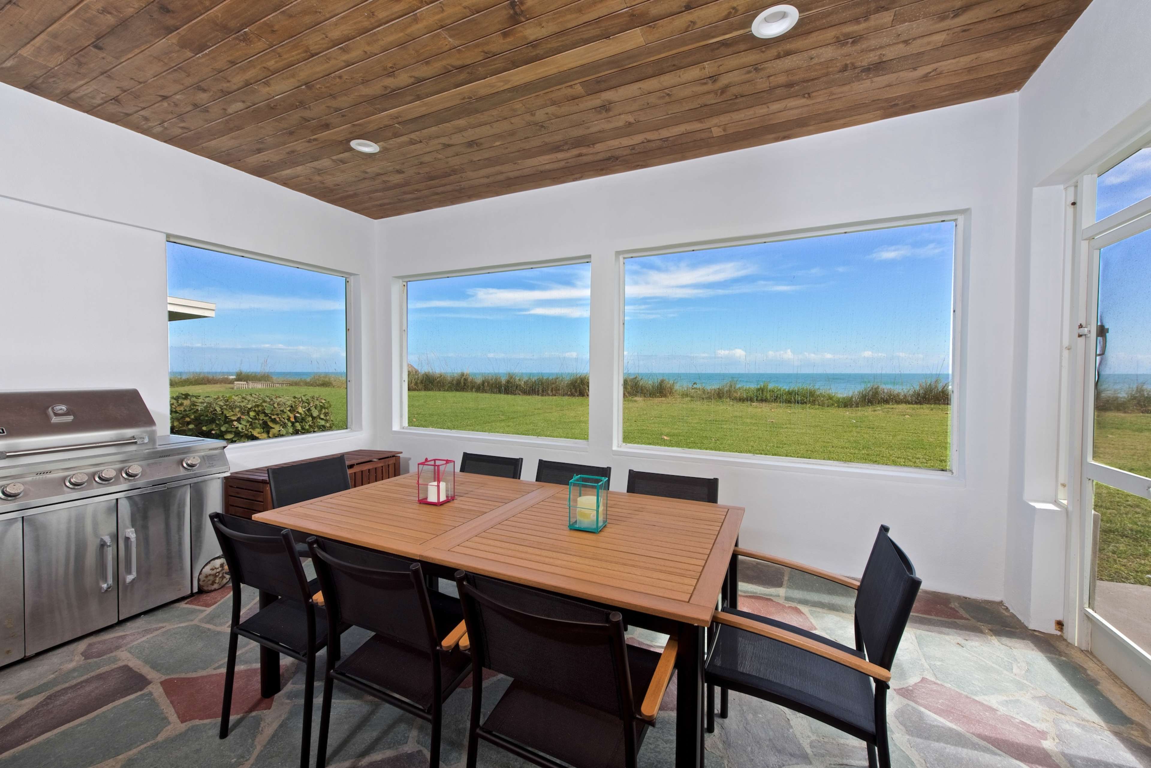 A spacious dining area features a wooden table surrounded by black chairs, with large windows showcasing a view of a grassy lawn and the ocean beyond.