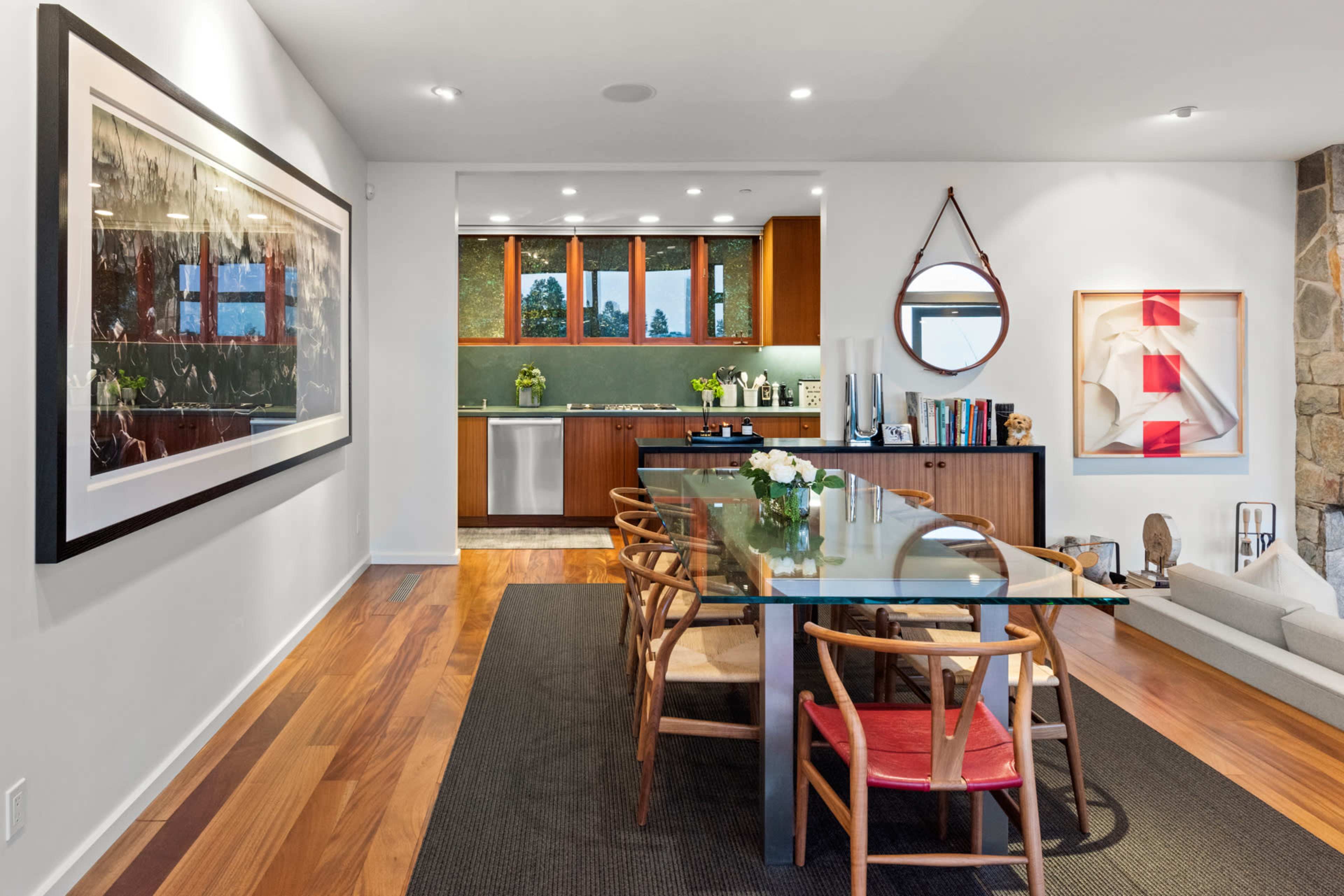 The image shows a modern dining area with a long glass table surrounded by wooden chairs, leading to a kitchen with wooden cabinetry and green walls.