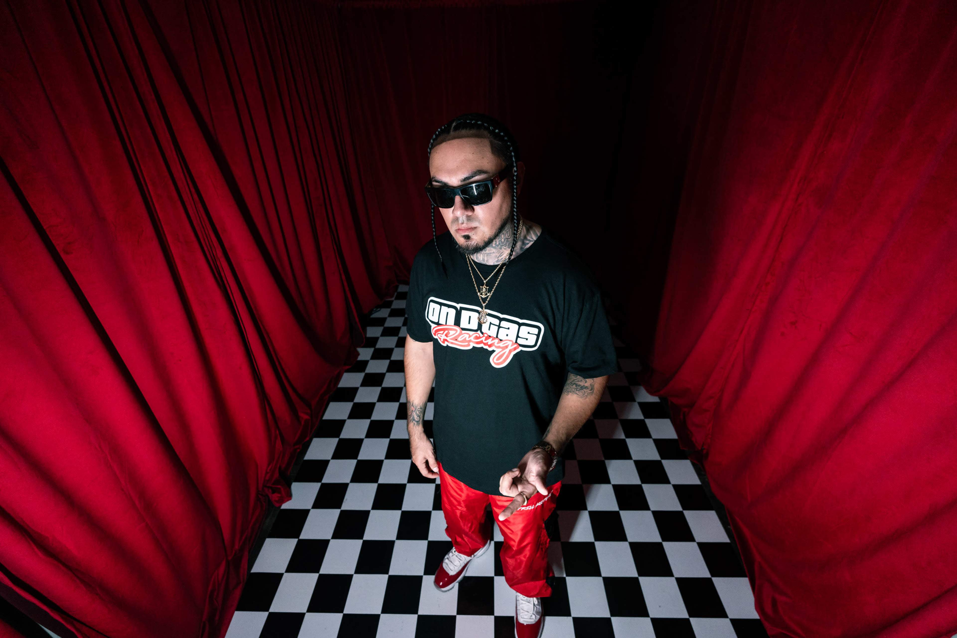 A person wearing sunglasses and a black shirt stands on a black-and-white checkered floor surrounded by red curtains.