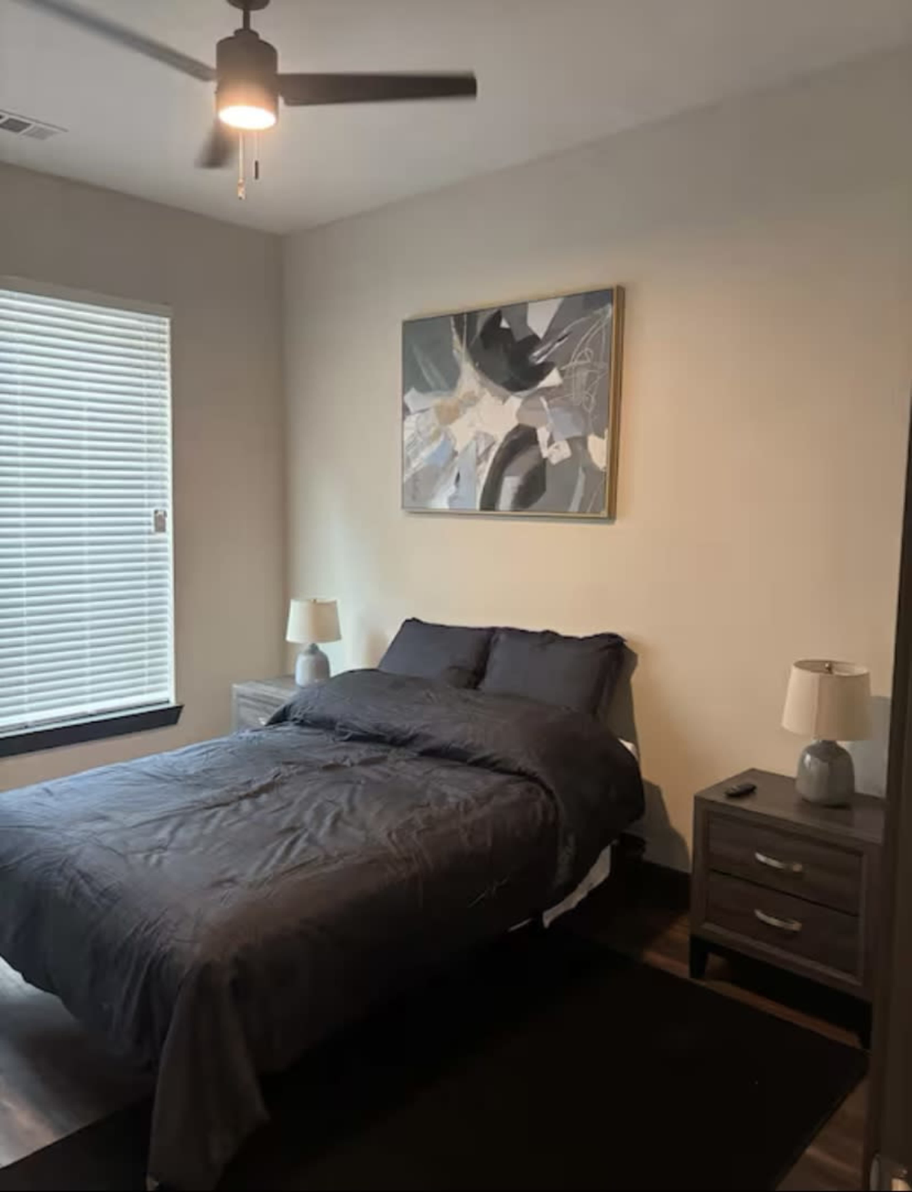 The image shows a bedroom featuring a bed with a dark-colored comforter, two nightstands with lamps, a window with blinds, and a wall-mounted painting.