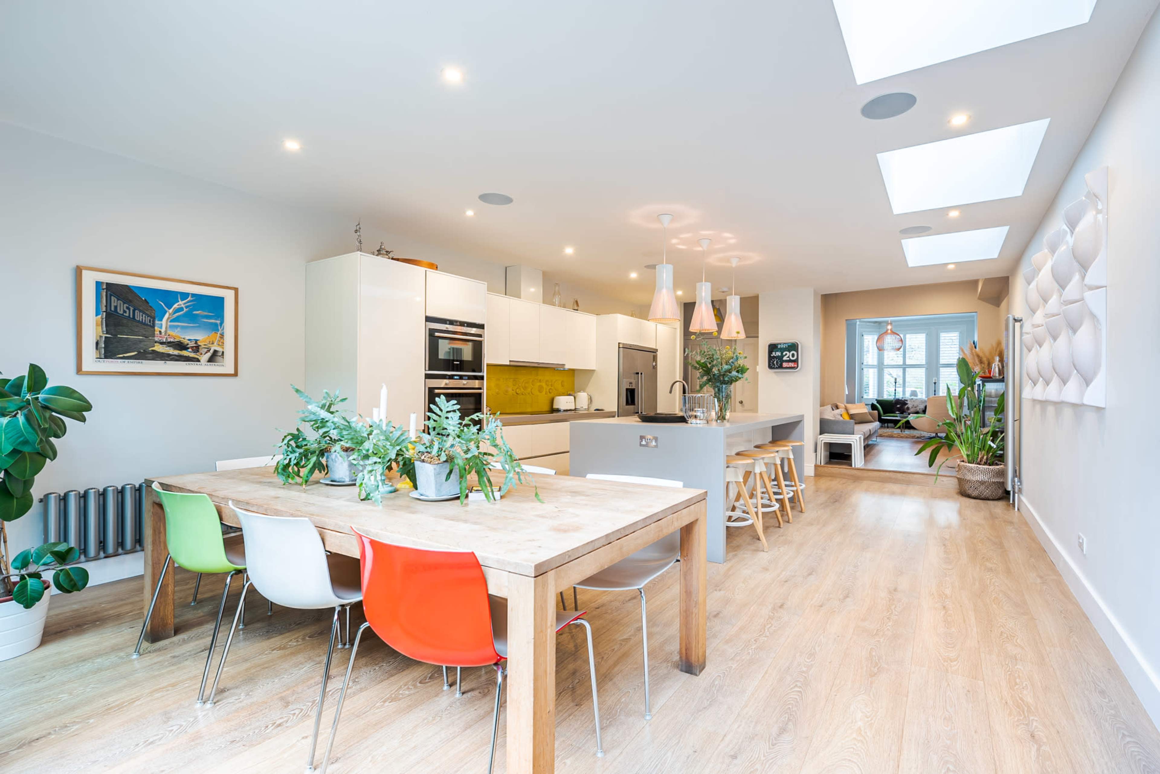 A spacious modern kitchen features a wooden dining table with colorful chairs, sleek appliances, and ample natural light from large windows.