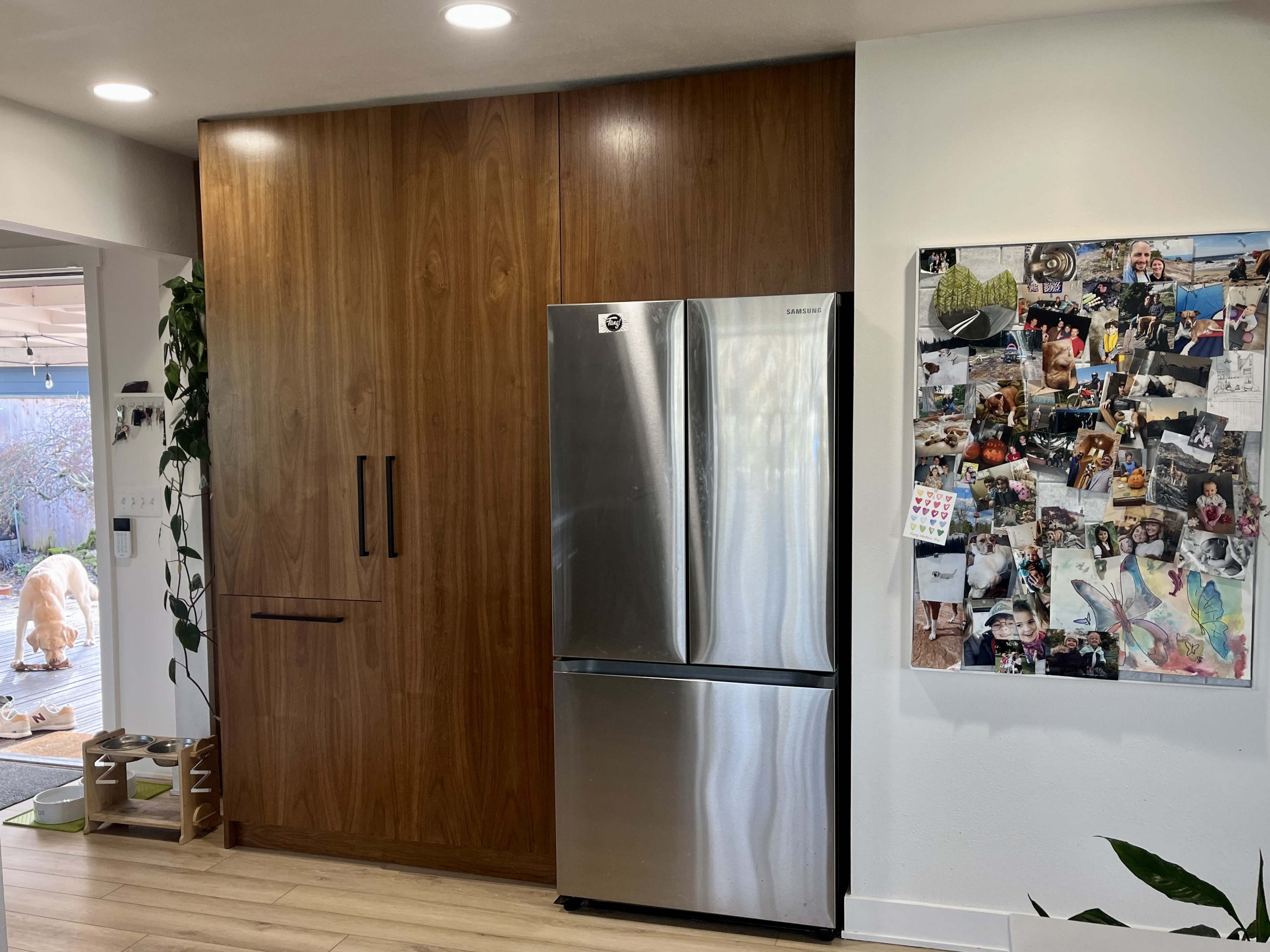 A modern kitchen with a stainless steel refrigerator next to a wooden cabinet, and a collage of photos mounted on a wall nearby.