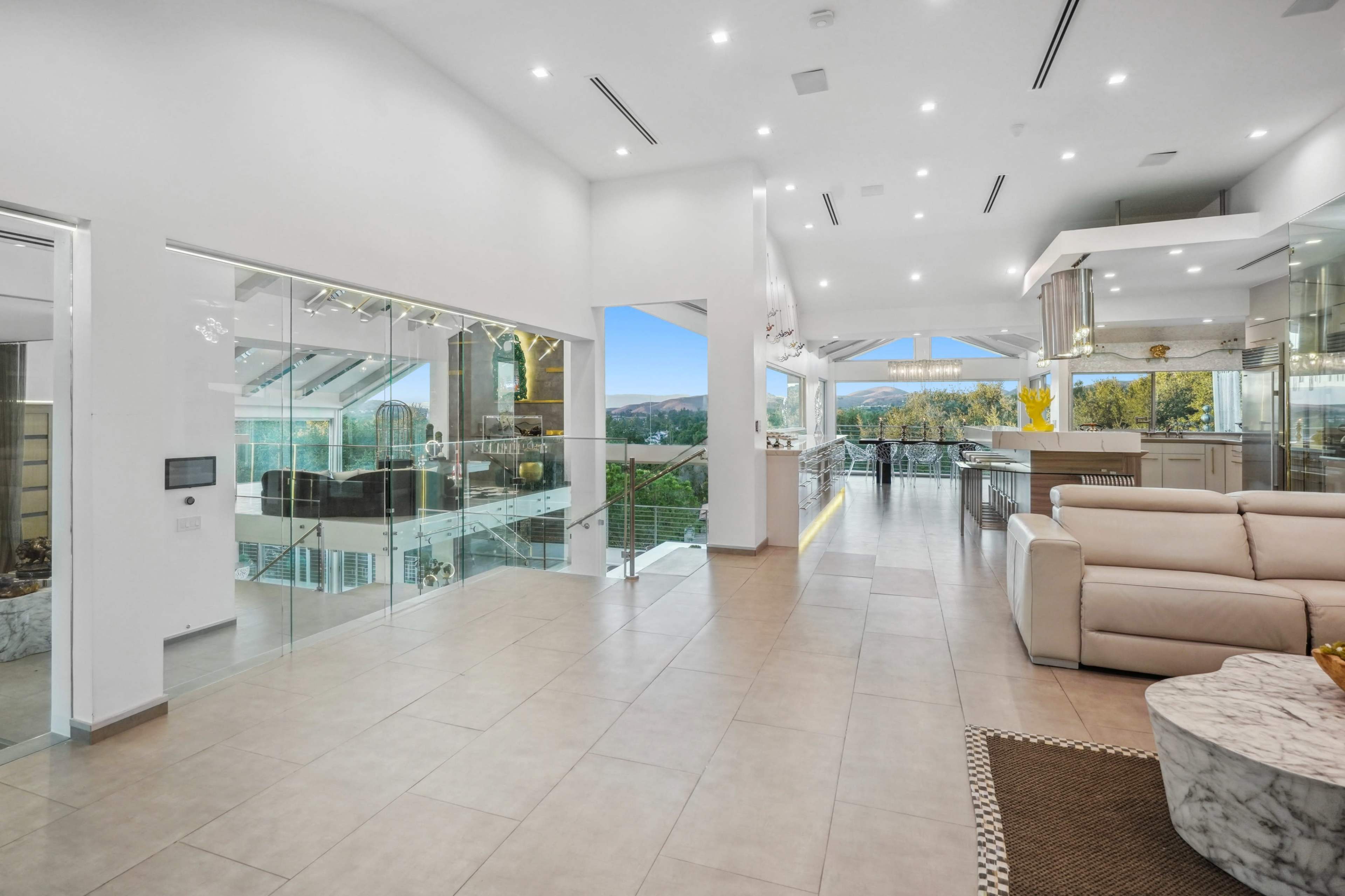 The image shows a modern, open-concept living space featuring large windows, a marble coffee table, and a sectional sofa, with a view of a landscape beyond.