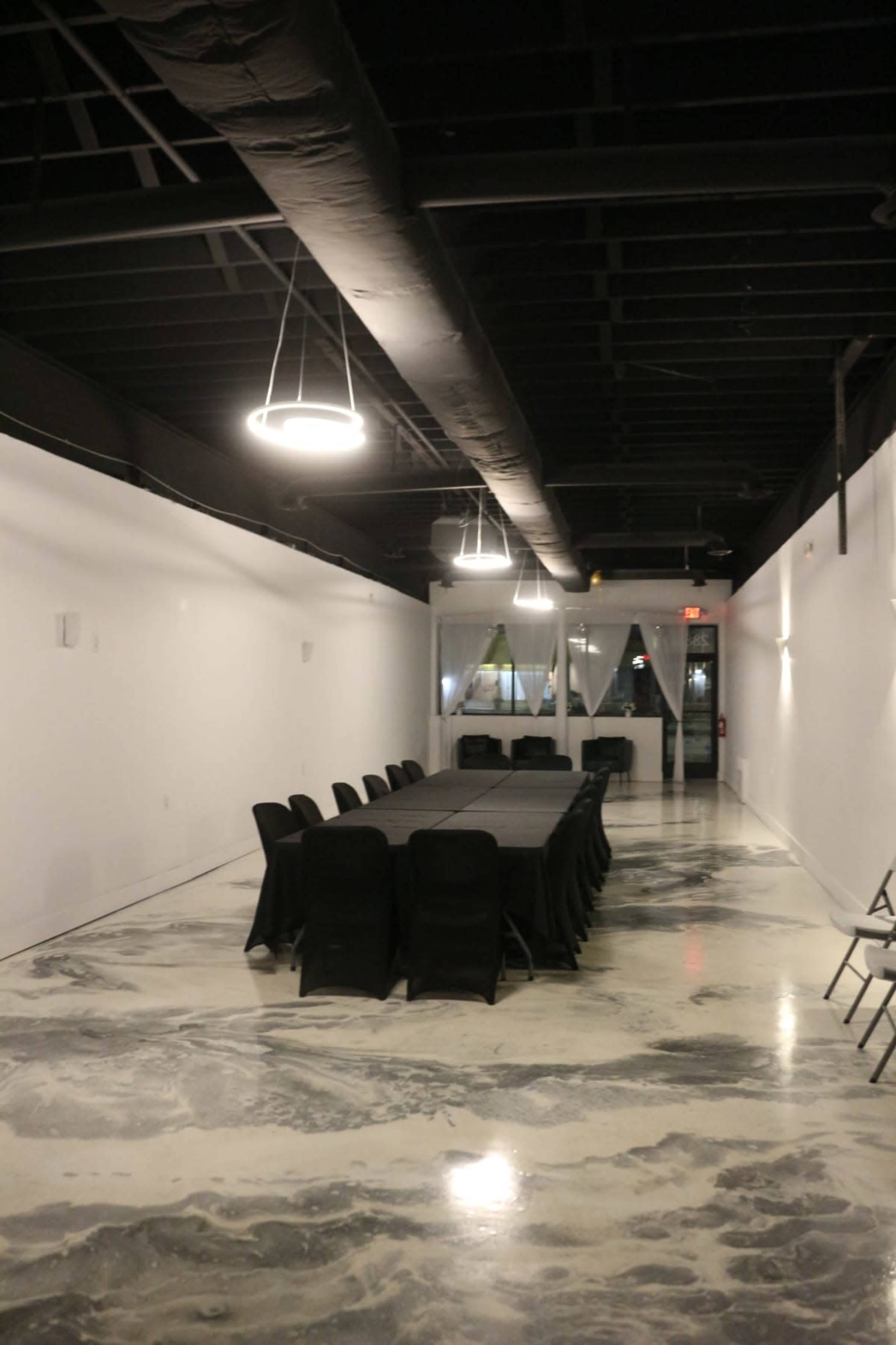 A long table with black chairs is set up in a spacious, minimally decorated room with concrete floors and overhead lighting.
