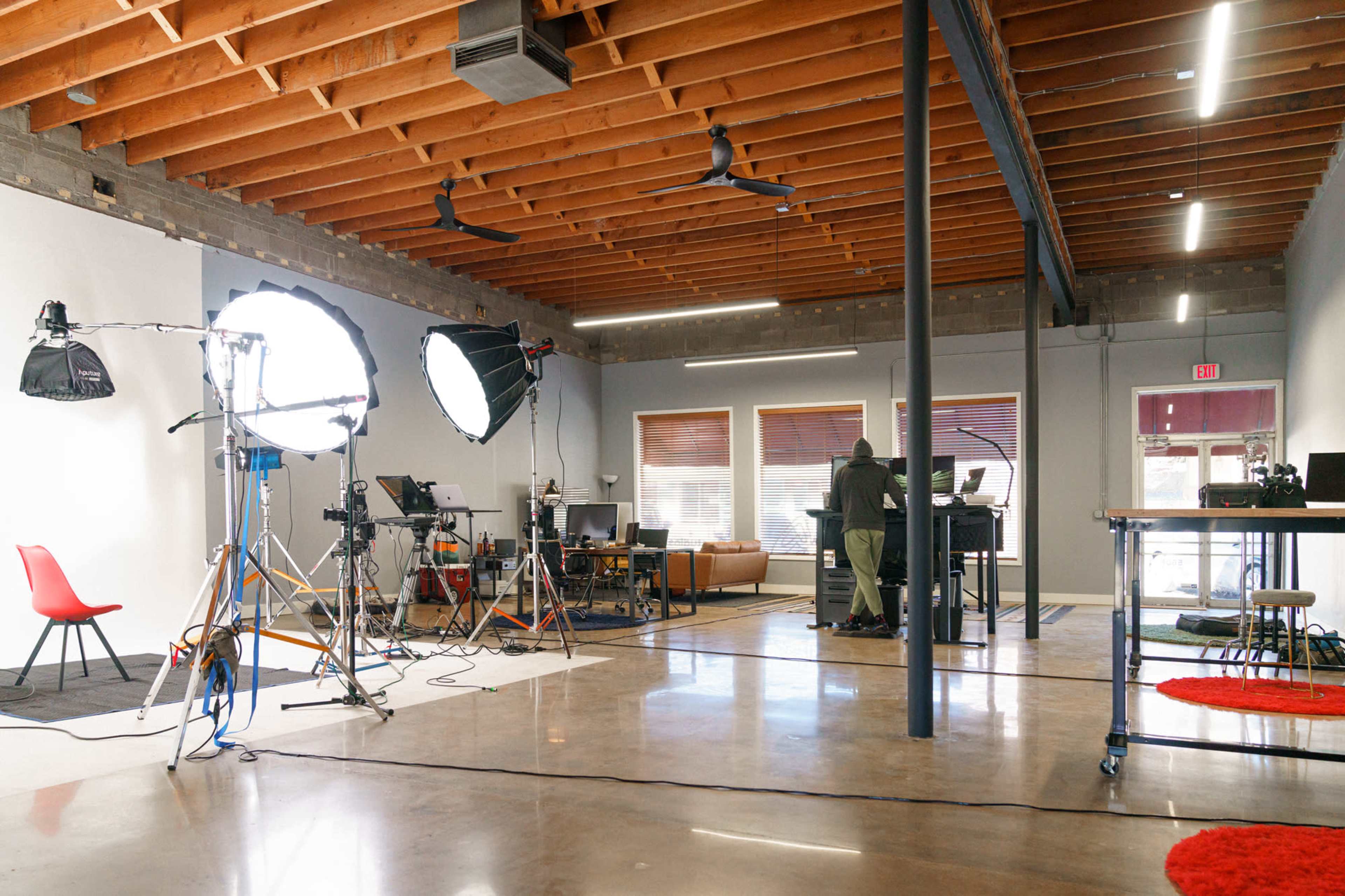 A spacious studio with various lighting setups and equipment, featuring a person working at a desk in the background.