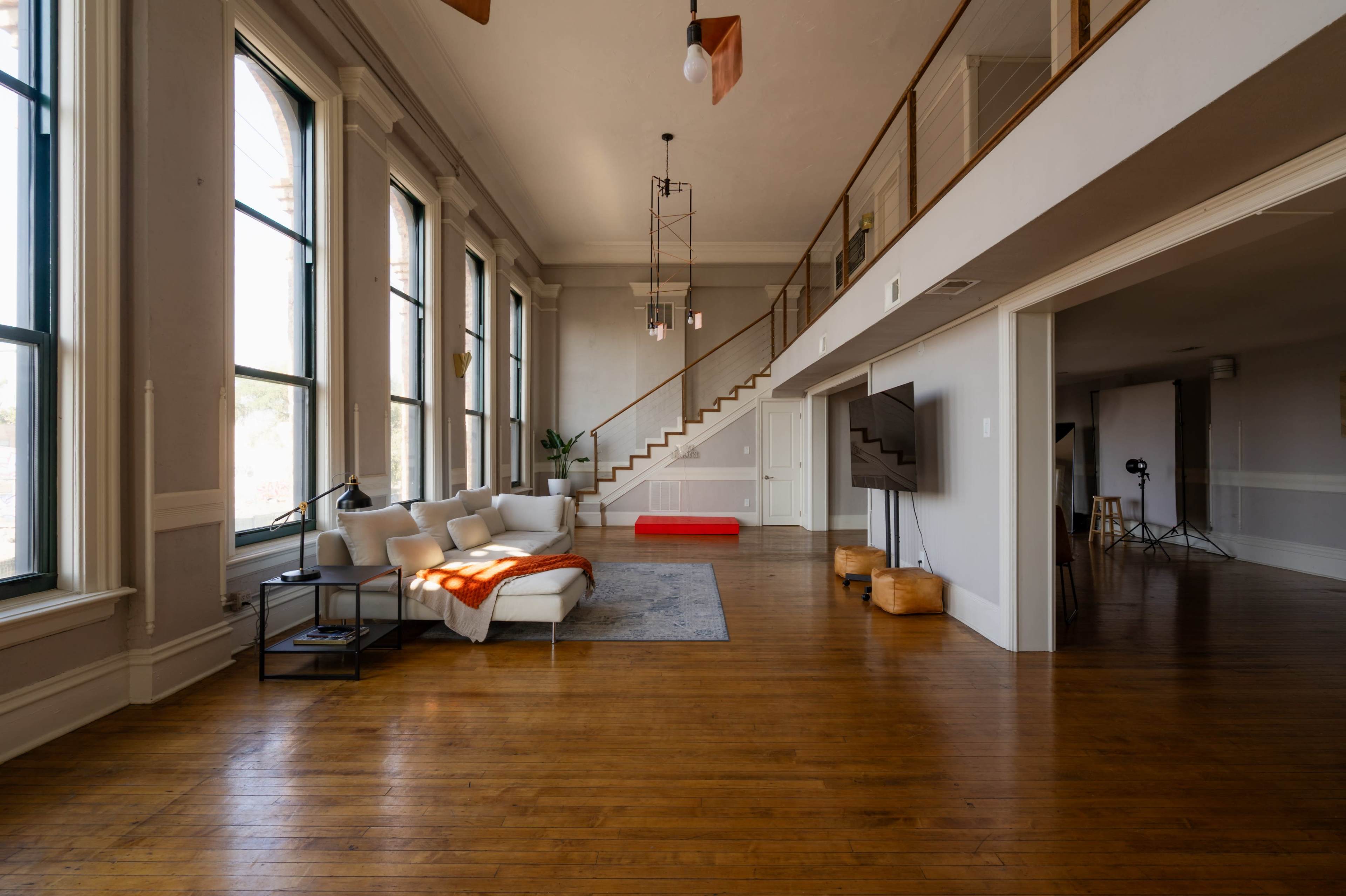 The image shows a spacious living area with hardwood floors, large windows, a modern sofa, and a staircase leading to an upper level.